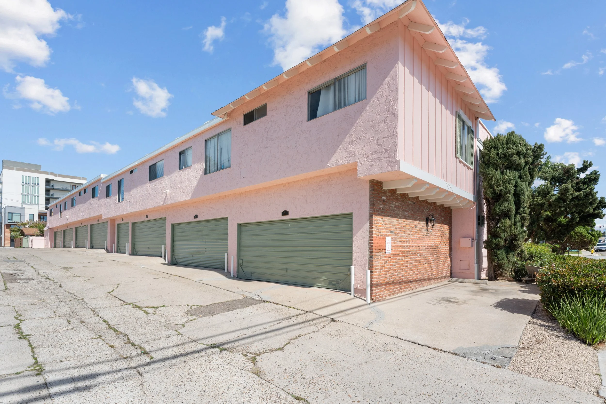 A pink stucco building with multiple garage doors at ground level, some windows, a brick section, and a small tree on the right side under a bright blue sky with scattered clouds.