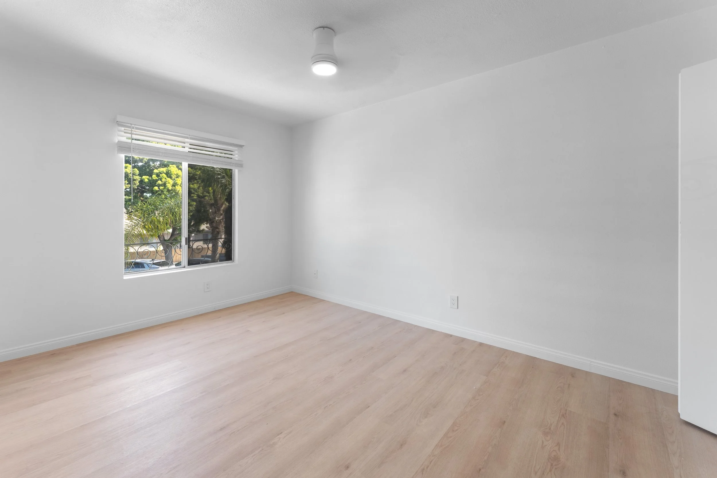 Empty room with white walls, light wood flooring, a window with view of trees and a balcony, and a ceiling light fixture.
