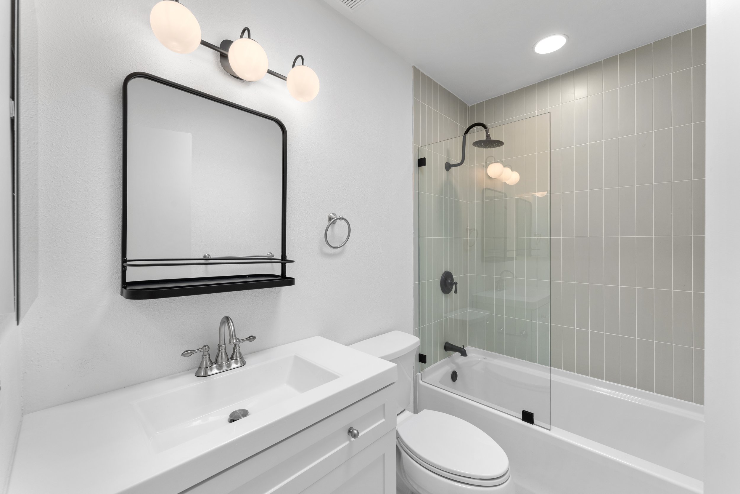 Modern bathroom with white vanity, black-framed mirror, wall-mounted light fixture, bathtub with glass shower enclosure, and beige tiled wall in the shower area.