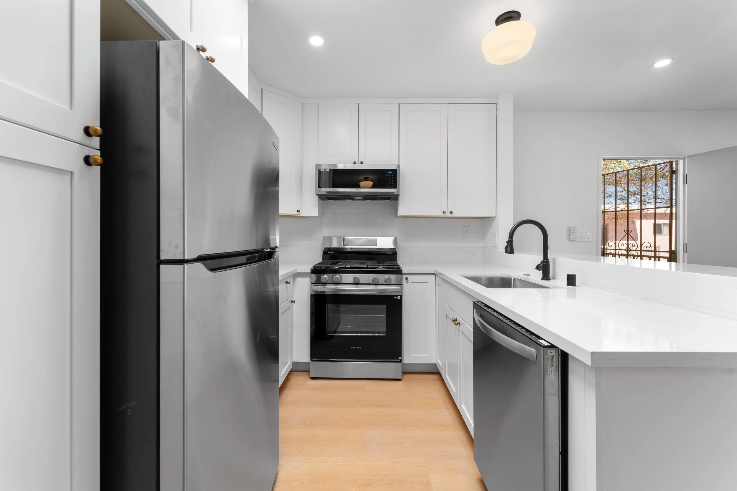Modern kitchen with white cabinets, stainless steel refrigerator, microwave, oven, and black faucet, hardwood flooring, and a window showing an outdoor view with trees.