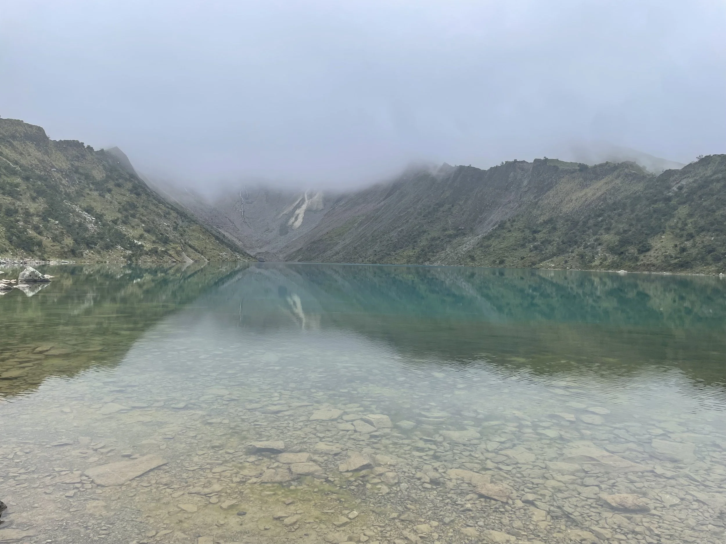 Paisaje de montaña reflejado en un lago en primer plano, rodeado de colinas verdes y neblina en el horizonte.
