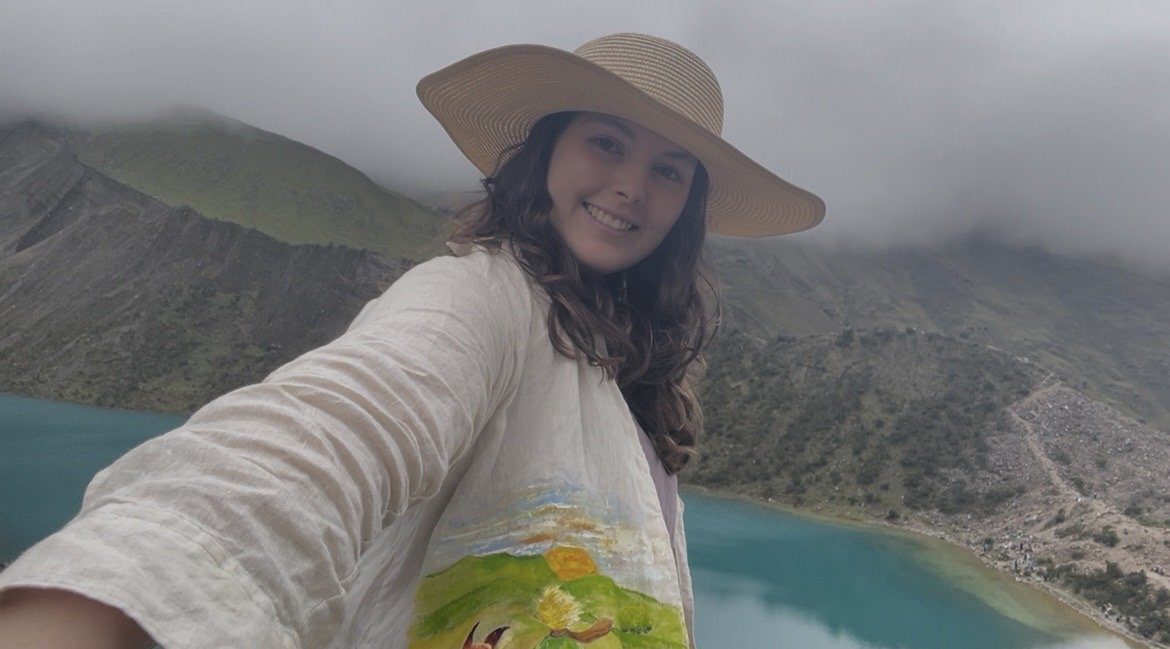 Joven mujer sonriendo, usando sombrero de ala ancha, con paisaje montañoso y lago en el fondo.