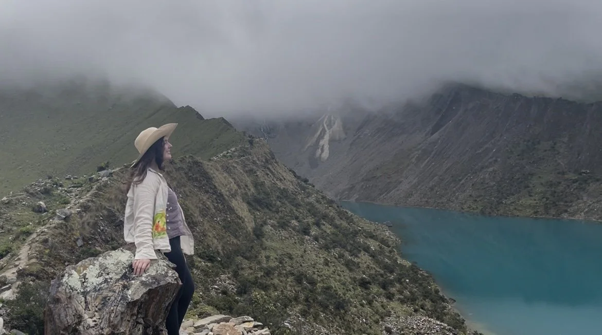 Mujer con sombrero de ala ancha en un paisaje montañoso con lago celeste y nubes bajas.