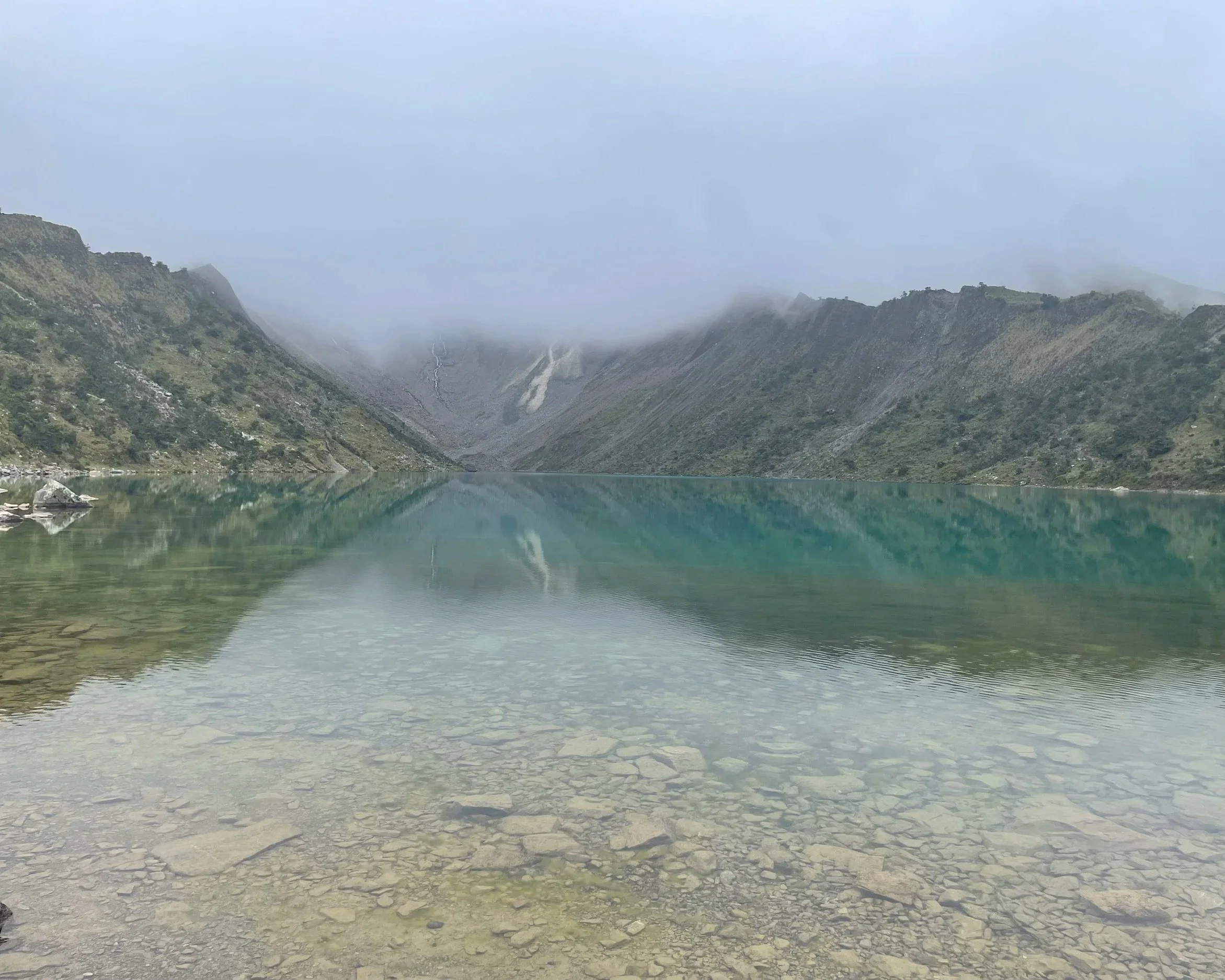 Un lago rodeado de montañas reflejadas en el agua con cumbres nubladas y vegetación verde, vista desde la orilla con agua clara en primer plano.