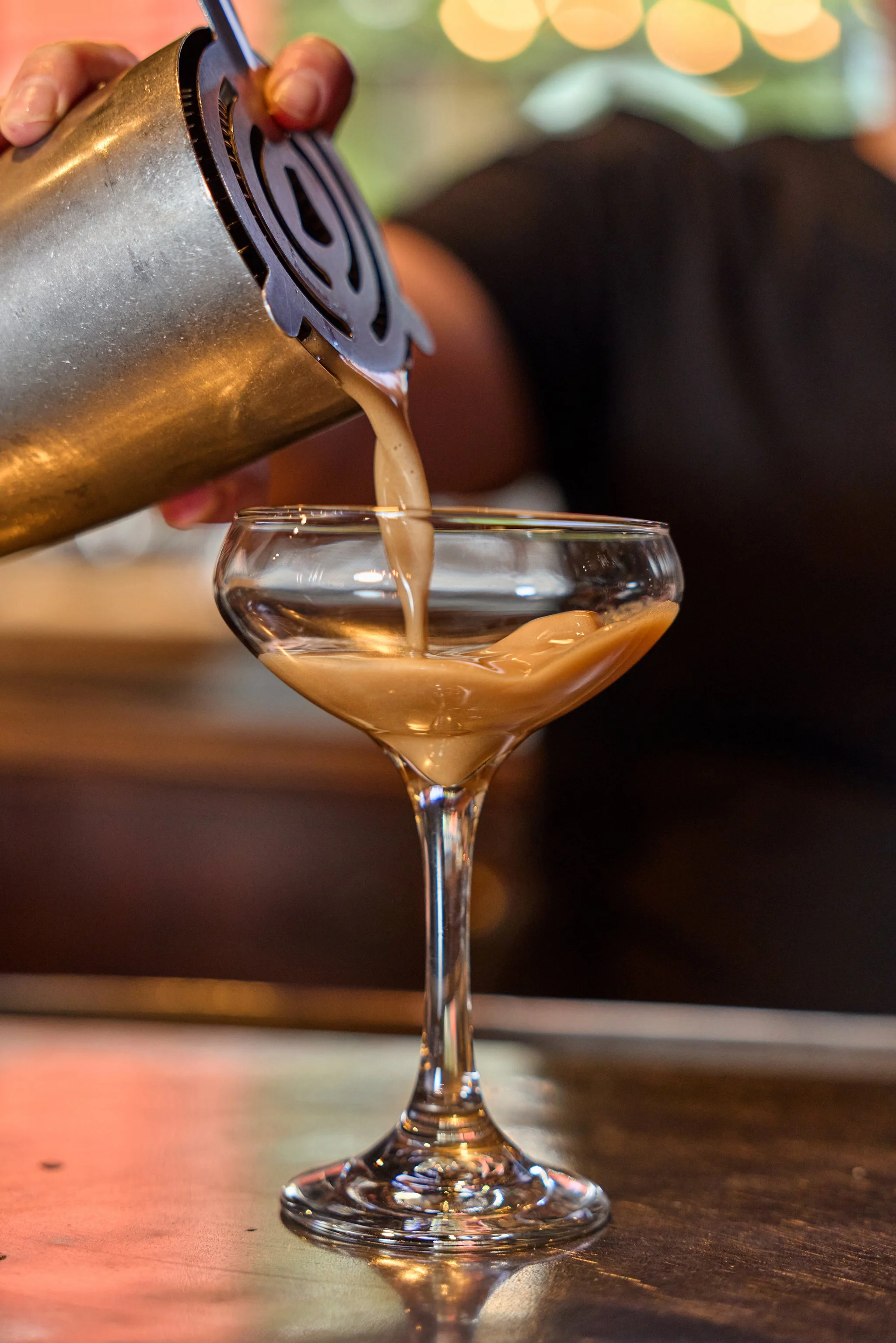 Pouring a creamy, light brown cocktail from a shaker into a martini glass on a wooden surface with blurred colorful lights in the background.