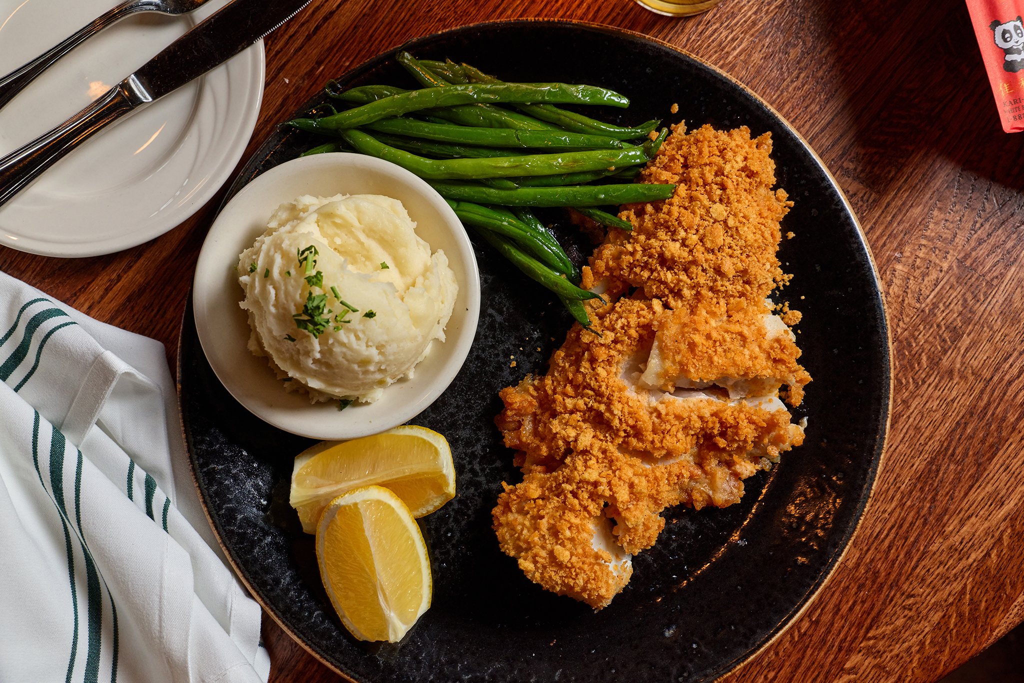 Plate of fried fish fillets with breading, green beans, mashed potatoes garnished with parsley, and lemon wedges on a black plate on a wooden table.