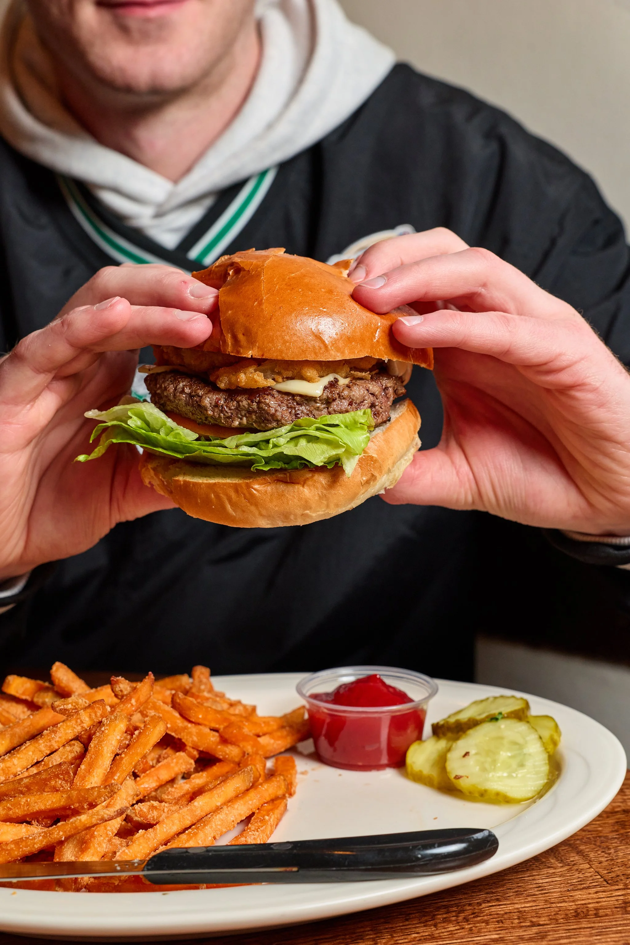 A person holding a cheeseburger with lettuce, tomato, and onion, with French fries, pickles, and ketchup on a plate in the foreground.