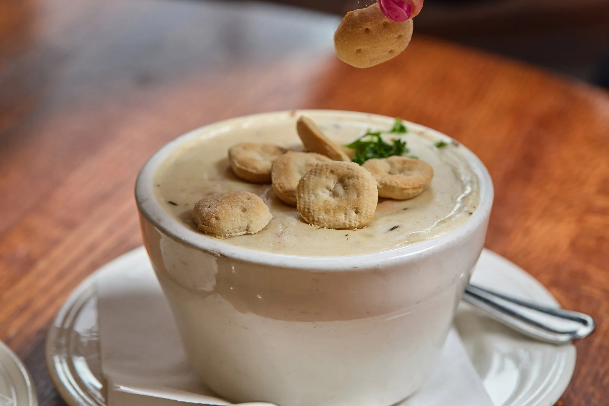 A cup of creamy soup topped with small crackers and garnished with chopped herbs, with a spoon placed beside on a plate.