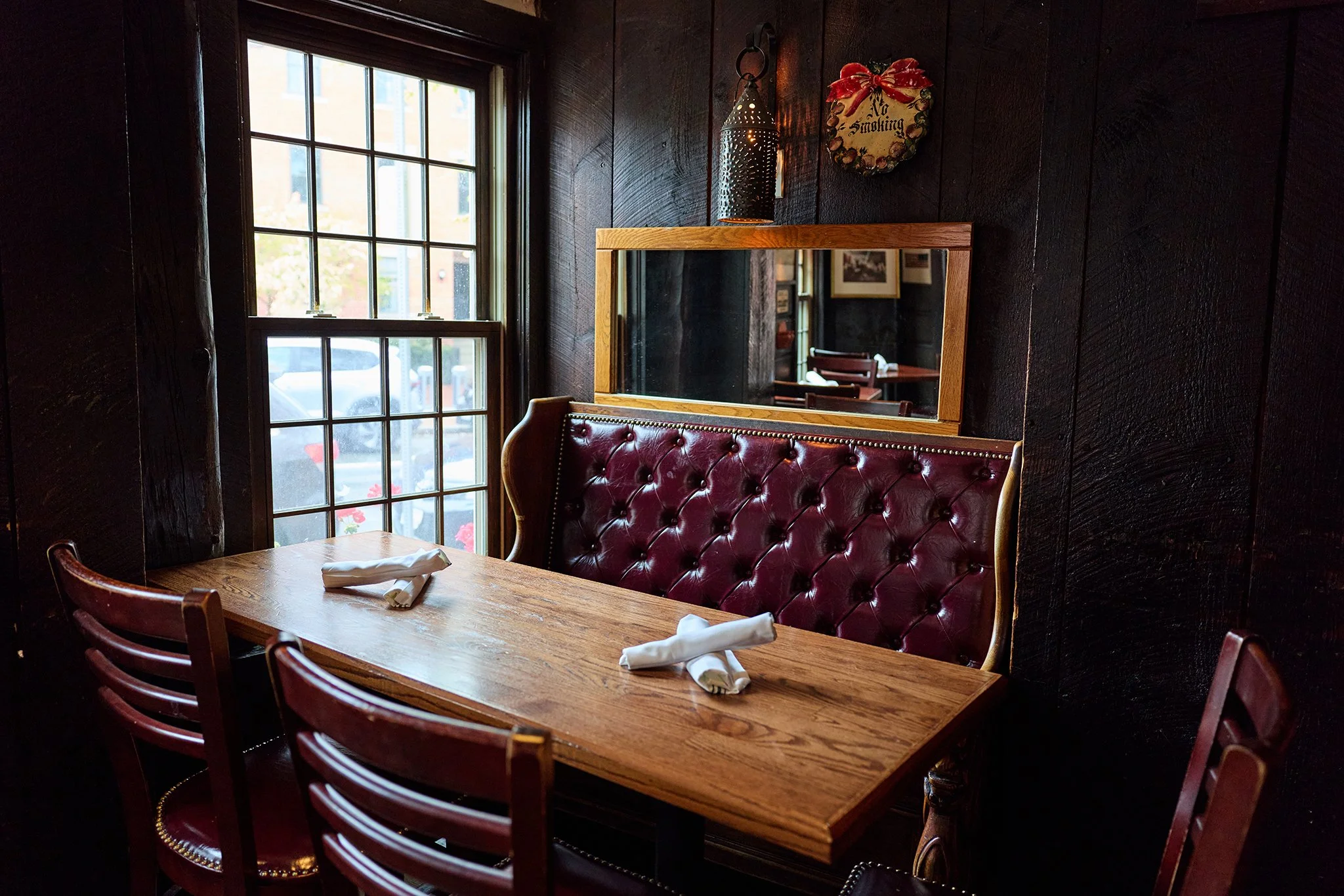 Inside a cozy restaurant with a dark wooden wall, a large window with multiple panes, a wooden table set with napkins, a tufted red leather bench, a mirror, and a wall decoration with a 