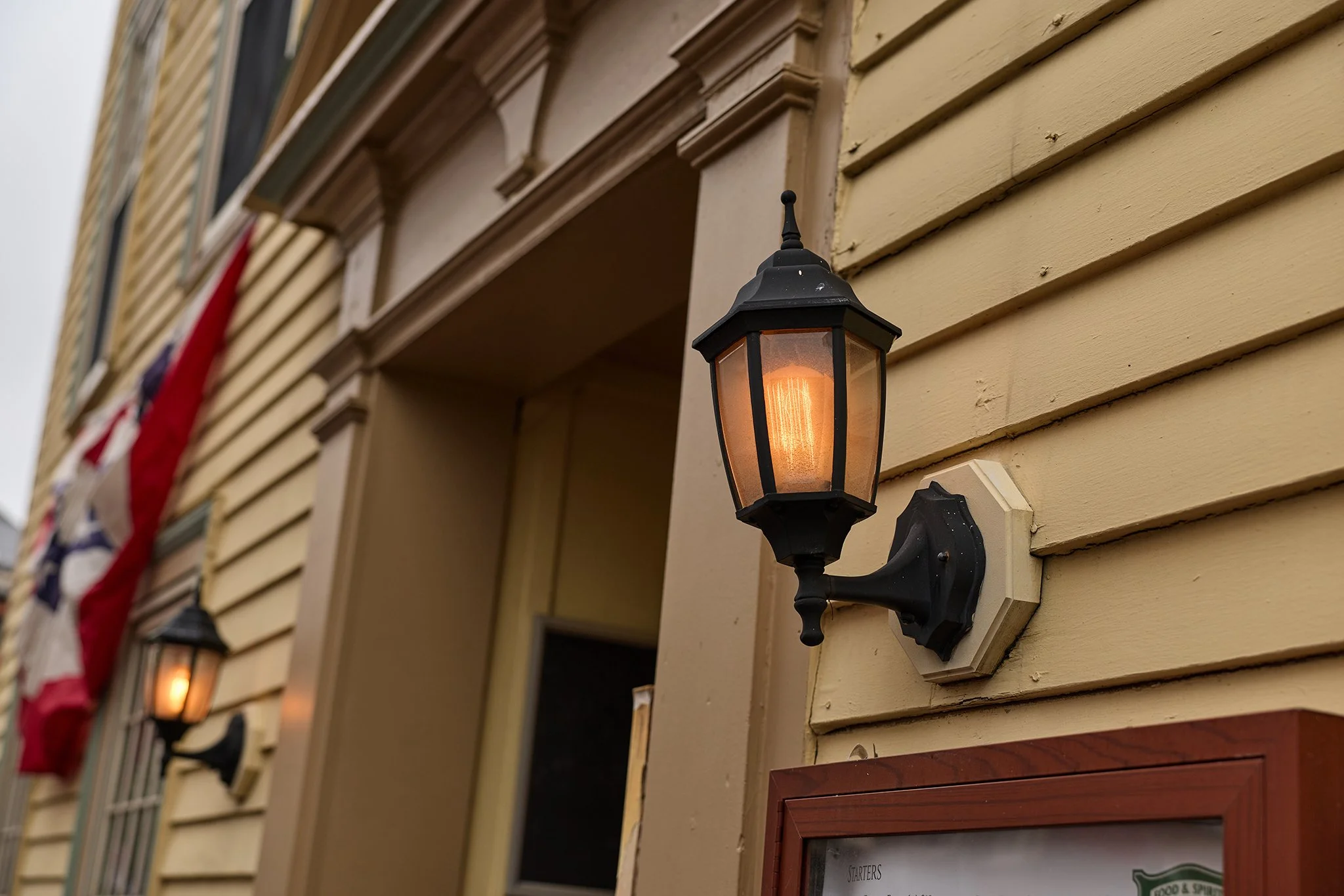 Exterior wall with beige horizontal siding, decorative trim, and black outdoor wall lantern with lit bulb.