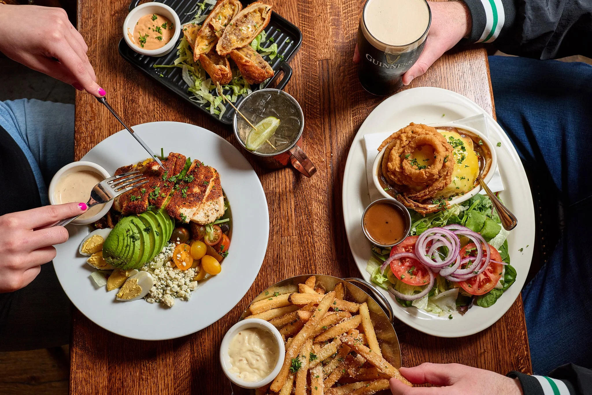 Table with various dishes including salads, fried foods, and drinks, with people reaching for food and a pint of Guinness beer.