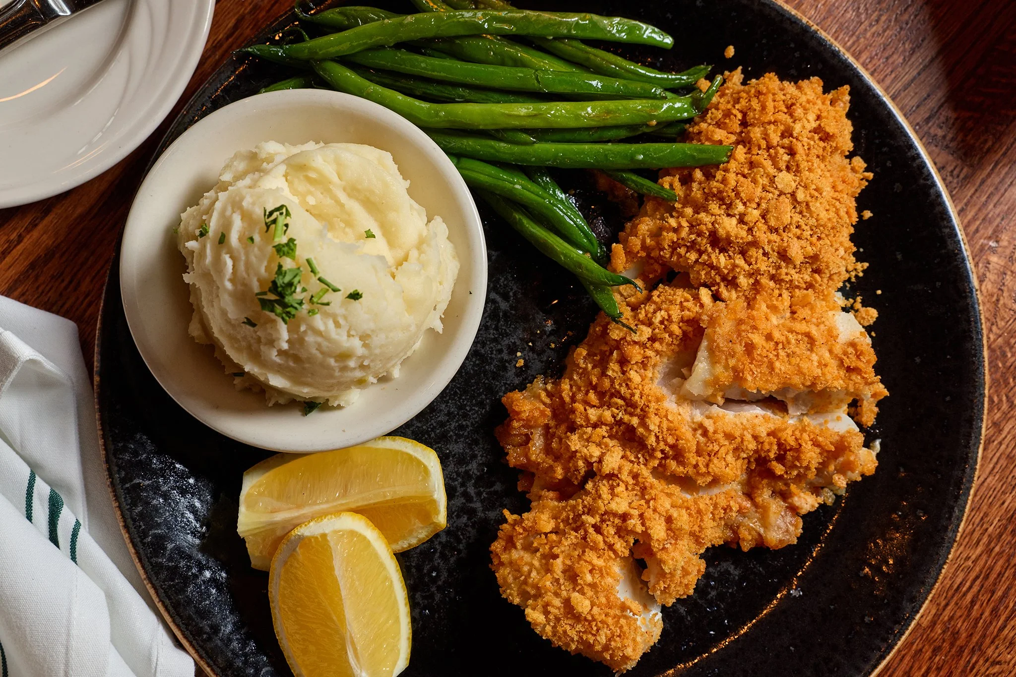 Fried fish fillet with breading, mashed potatoes with green herbs, green beans, and lemon wedges on a black plate.