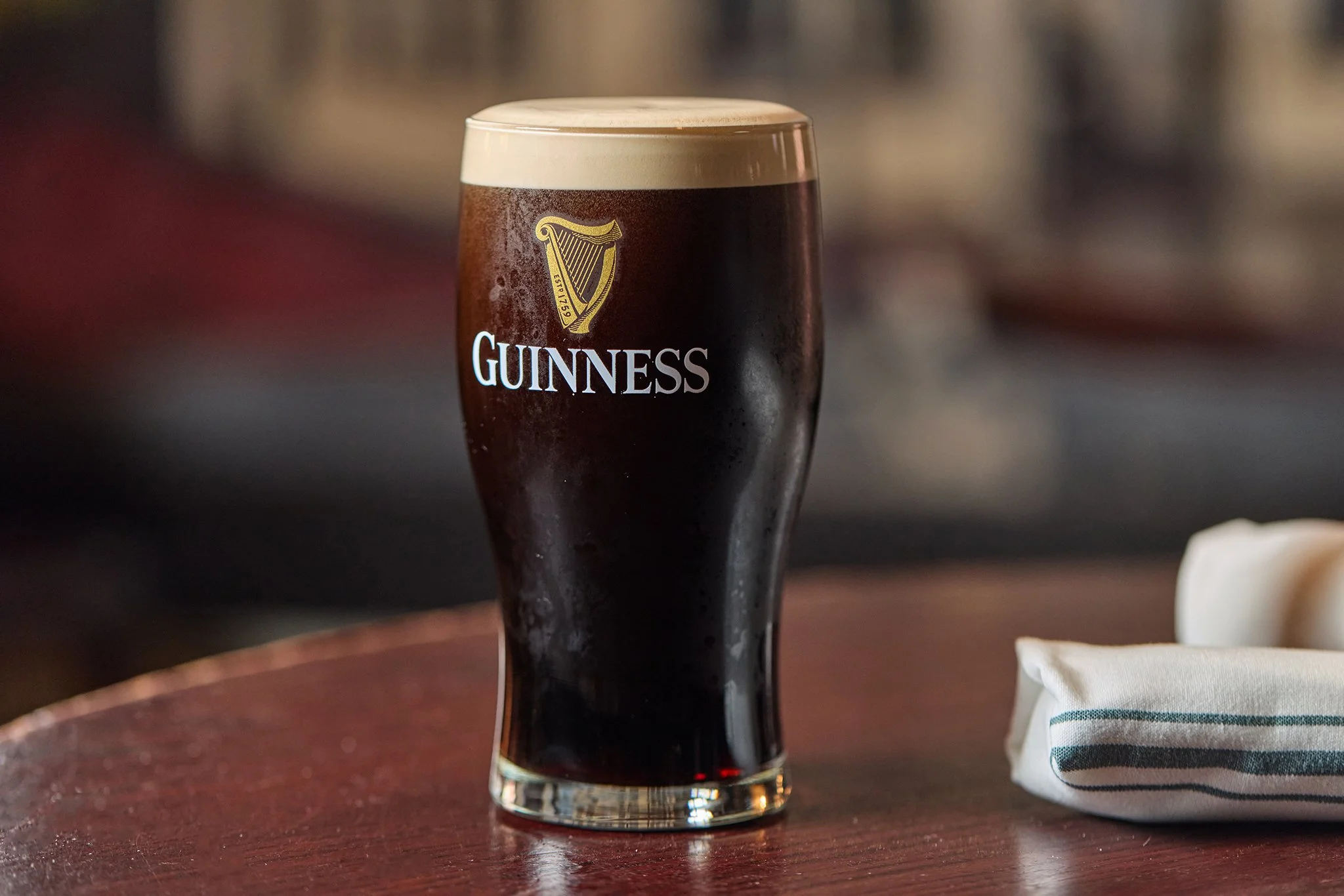 A glass of Guinness beer with a foamy head placed on a wooden table near a striped cloth napkin.