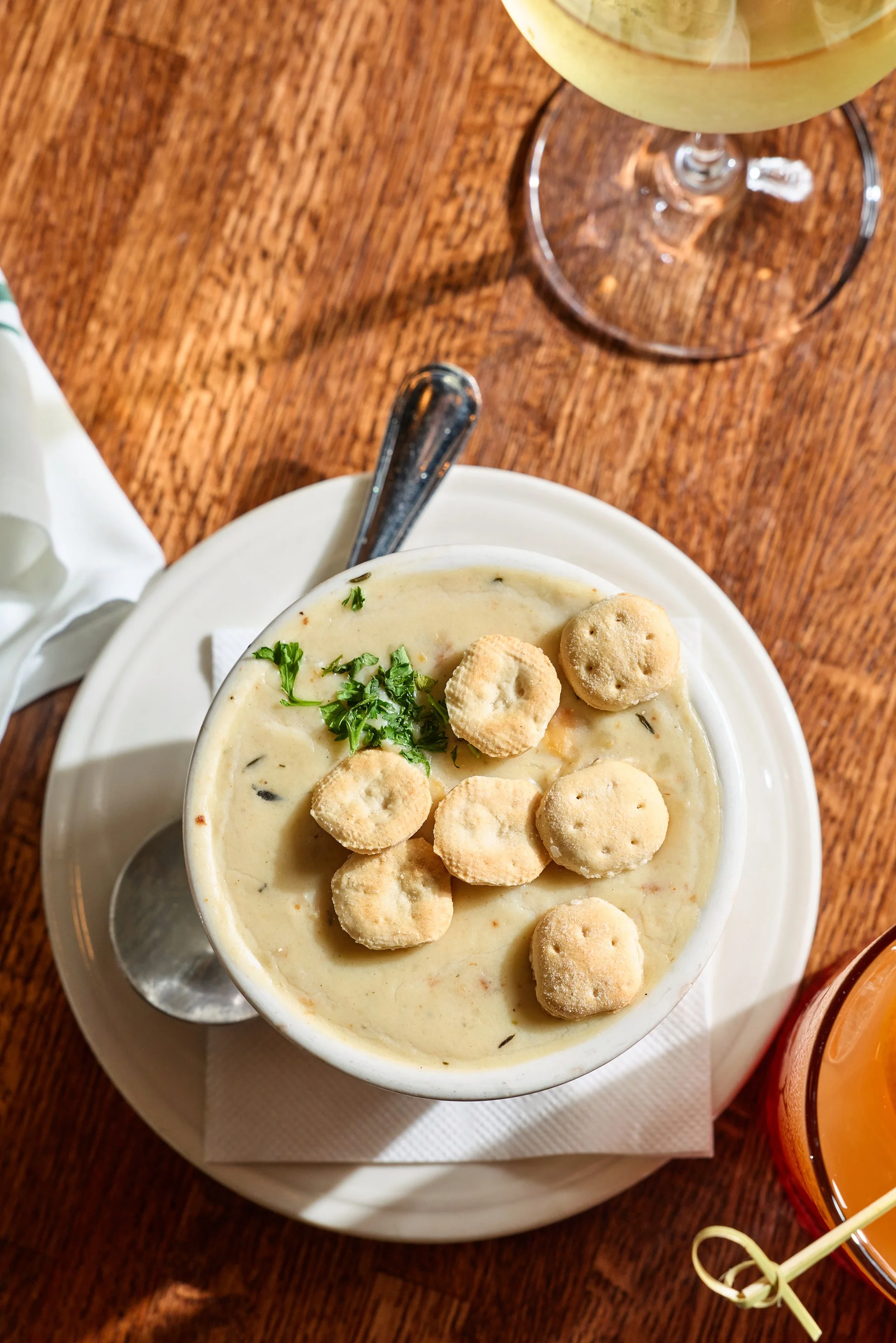 A bowl of creamy soup topped with small round crackers and chopped herbs, placed on a wooden table with a spoon inside the bowl. A glass of white wine and part of a glass of orange beverage are also visible.