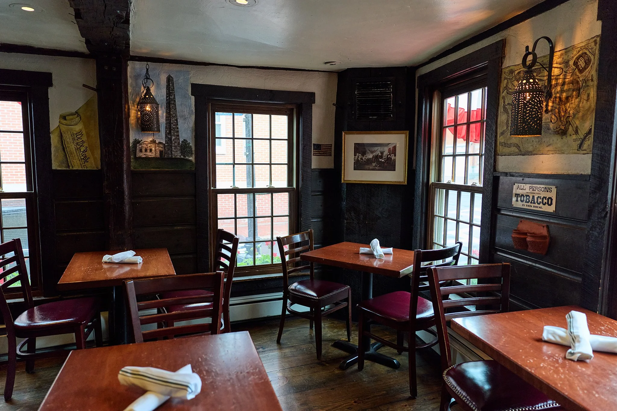 Interior of a cozy restaurant with dark wood furniture and large windows letting in natural light, decorated with vintage-style artwork and signs.