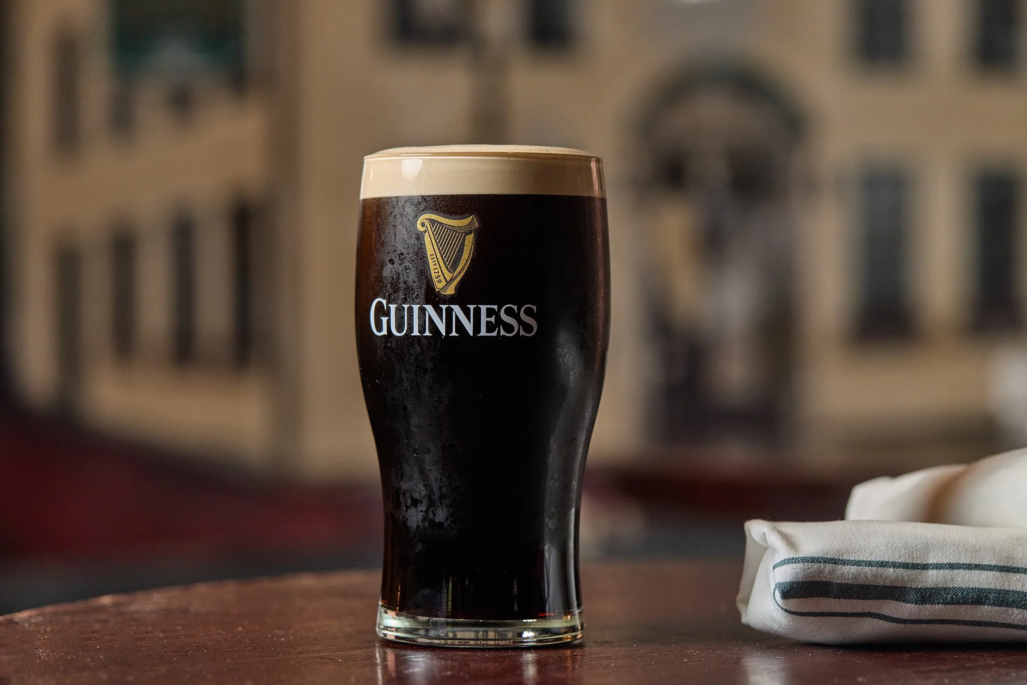 A glass of Guinness stout beer on a wooden table, with a blurred background and a cloth napkin on the right side.
