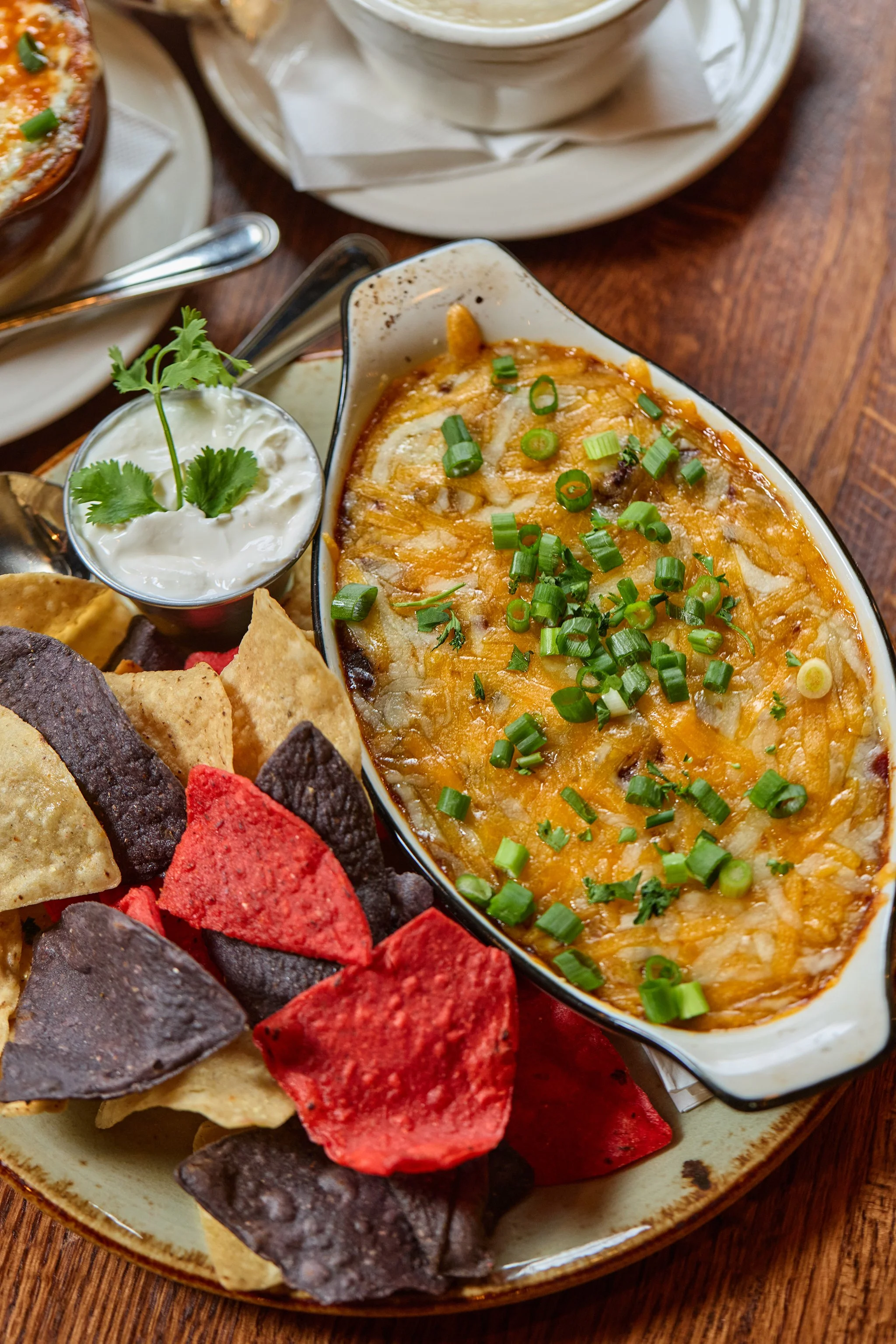 A serving of cheesy baked dip garnished with chopped green onions, accompanied by a small container of sour cream with cilantro, tortilla chips, and assorted colored chips on a plate, set on a wooden table.