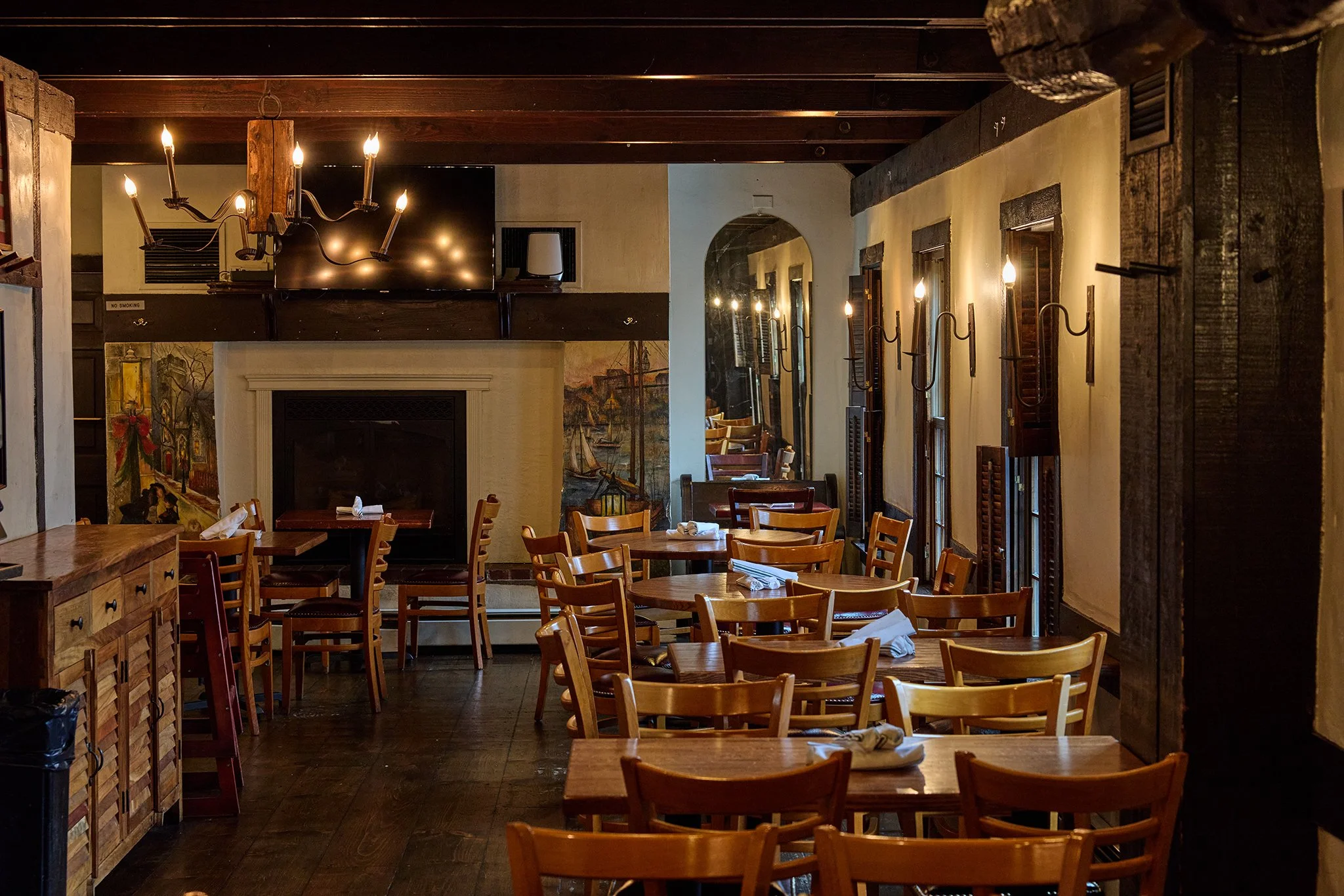 Interior of a cozy restaurant with wooden chairs and tables, warm ambient lighting, a fireplace, murals, and a dark wood ceiling.