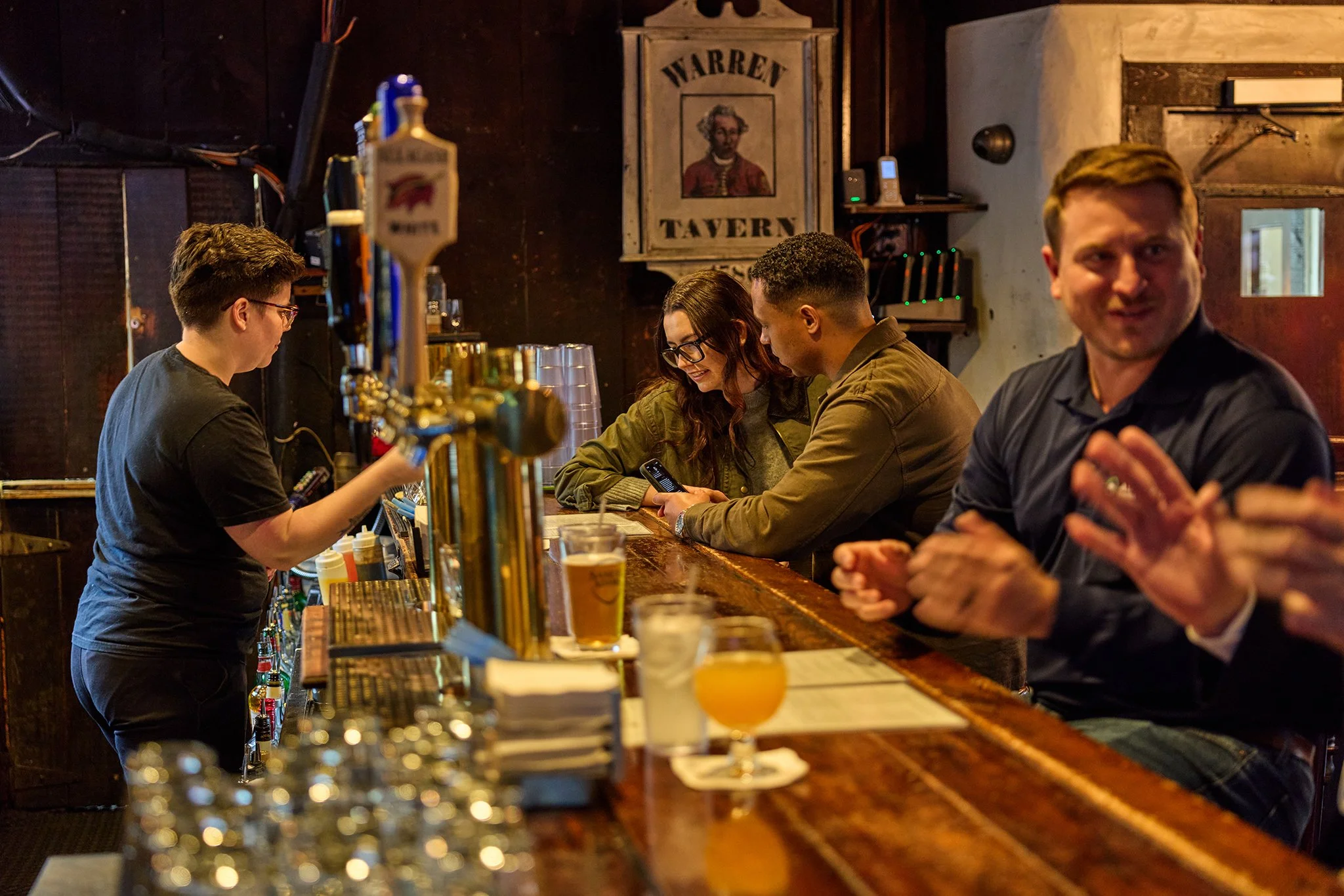 People sitting at the bar in a rustic tavern, including a bartender pouring a drink and a man smiling at the camera.