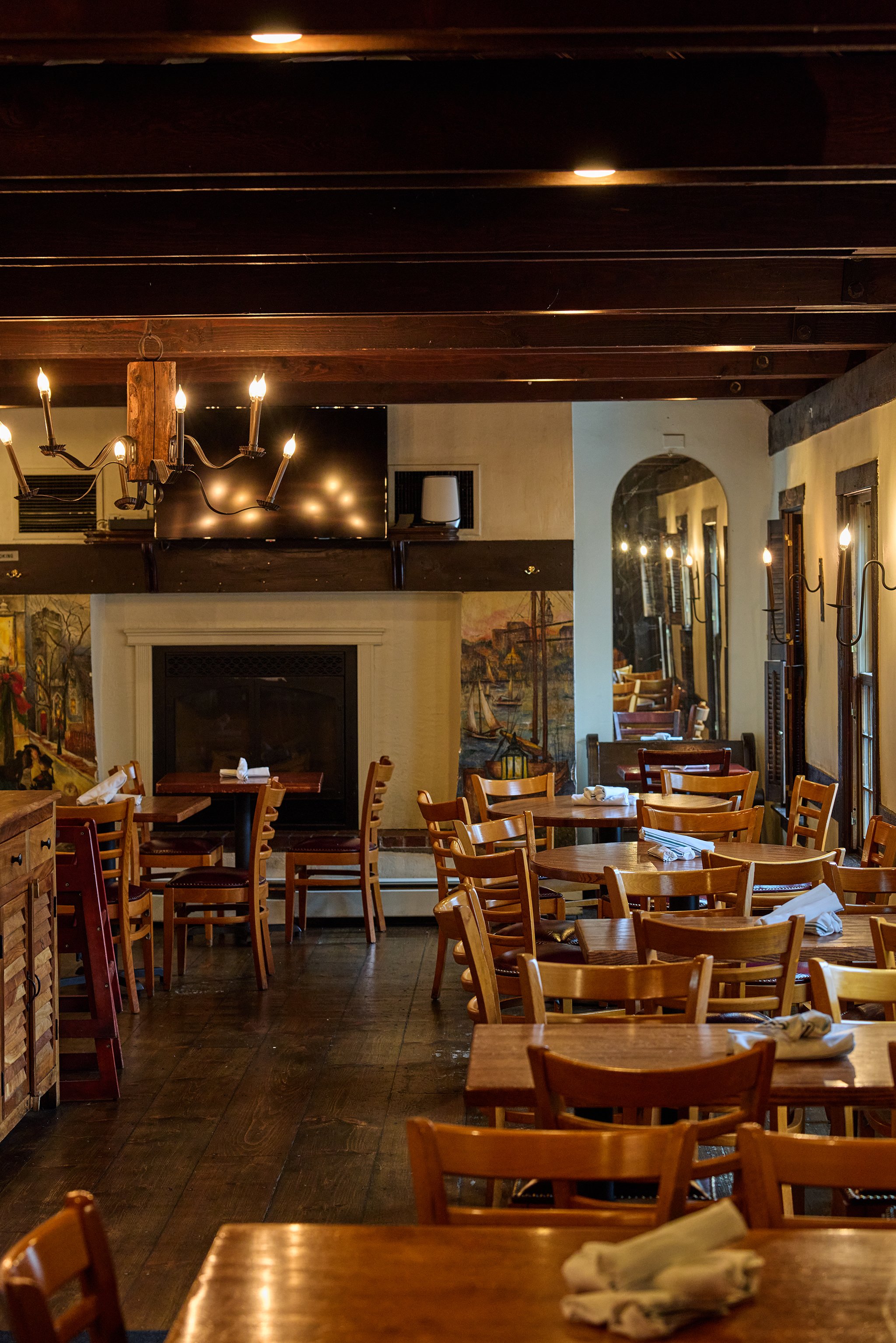 Interior of a cozy restaurant with wooden tables and chairs, a fireplace, paintings on the wall, and warm lighting.
