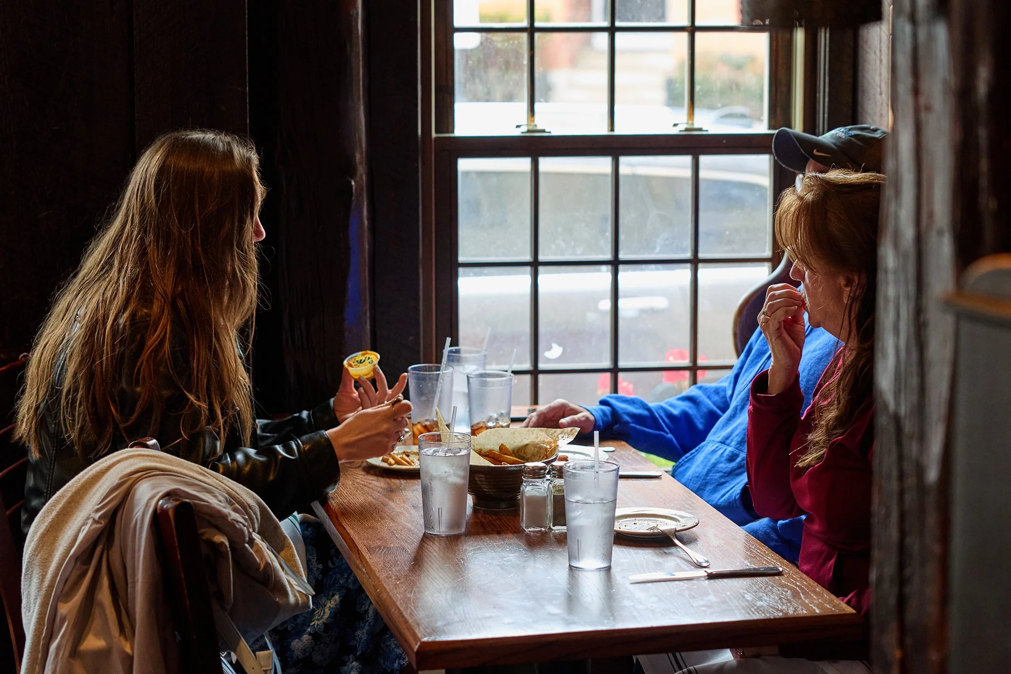 Two women sitting at a wooden table in a cozy restaurant, engaged in conversation, with food and drinks on the table and a large window behind them.