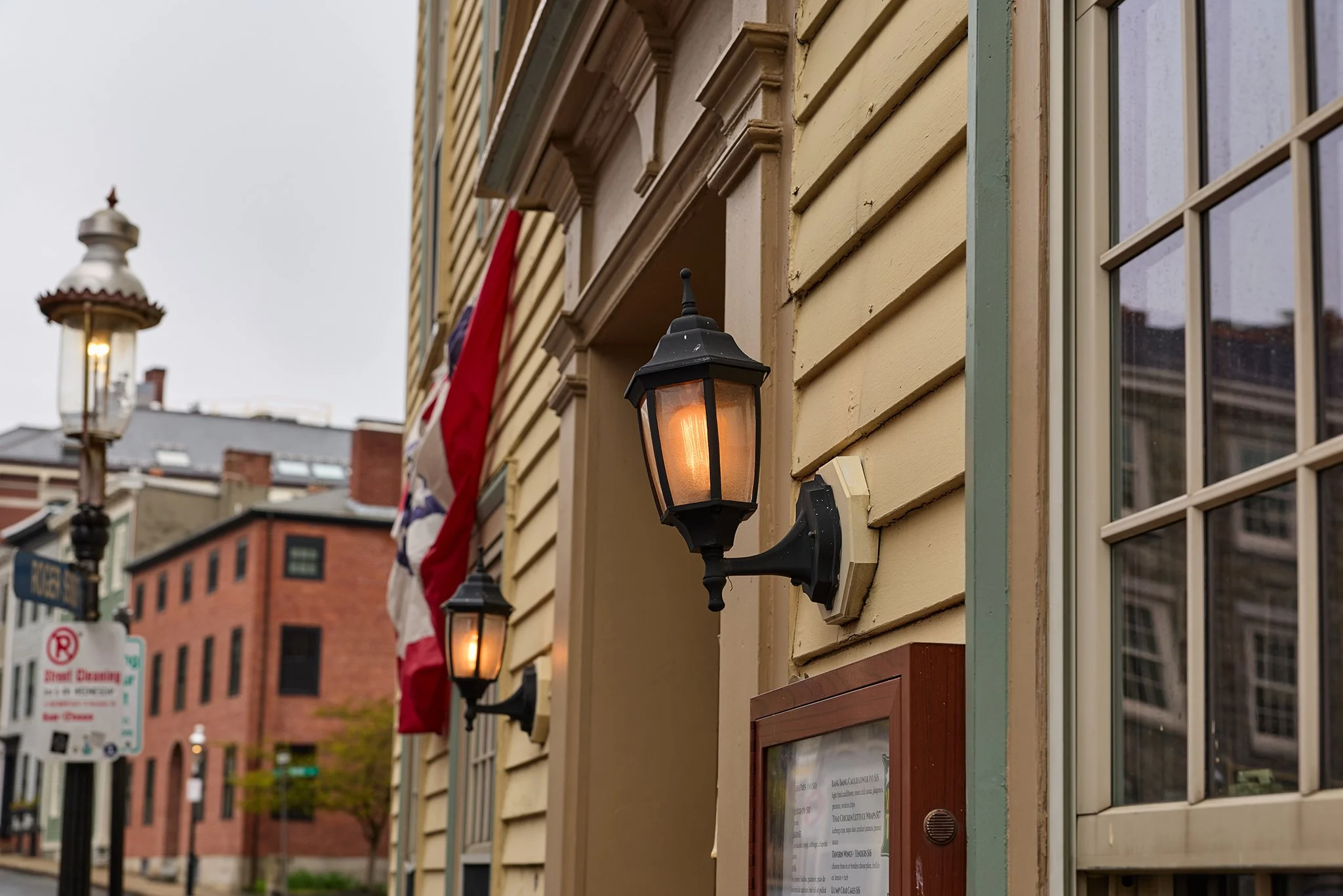 Close-up of an exterior wall with two black lantern-style lights, yellow wooden siding, a window, and flags hanging in the background. City street with buildings and a street sign.