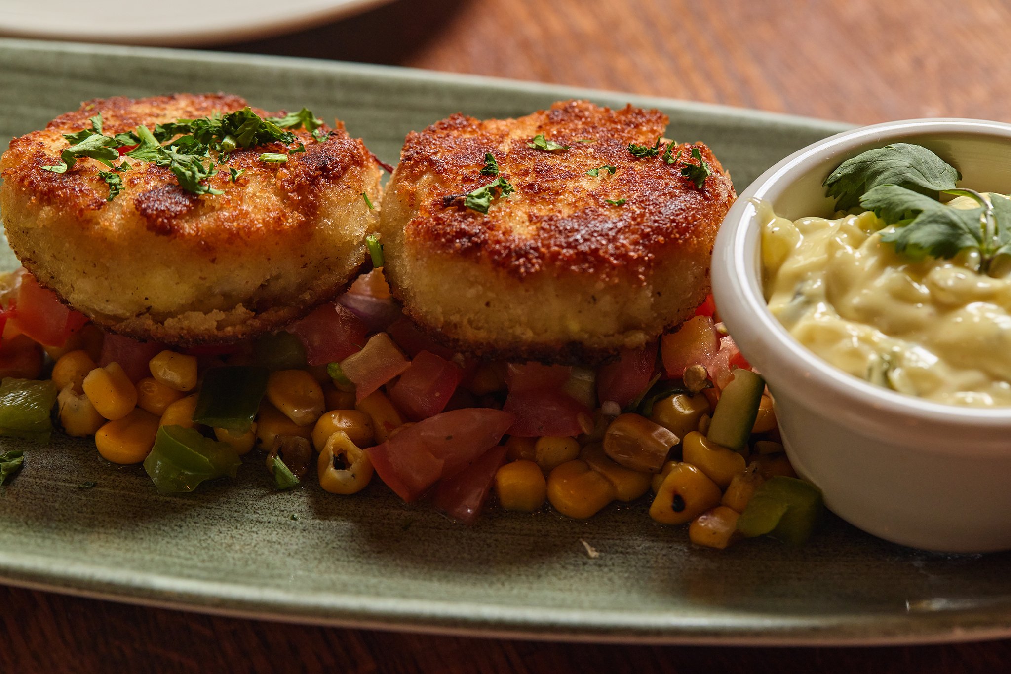 Two golden brown crab cakes garnished with chopped parsley, served on a bed of mixed vegetables with a side of creamy dipping sauce garnished with parsley, on a green ceramic plate.
