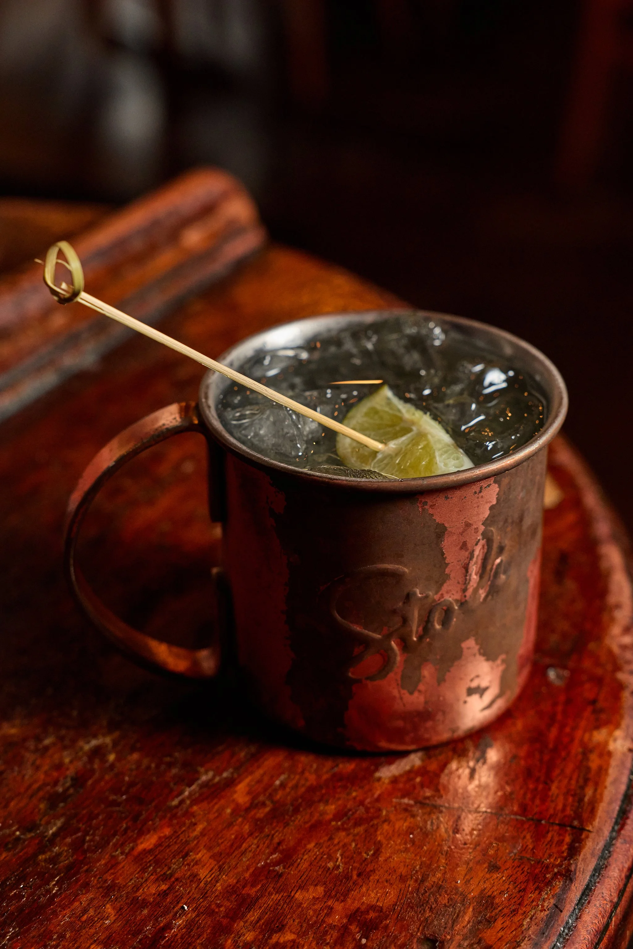 A metal cup with ice and a lime wedge, placed on a wooden surface, with a drink pick resting on the cup.