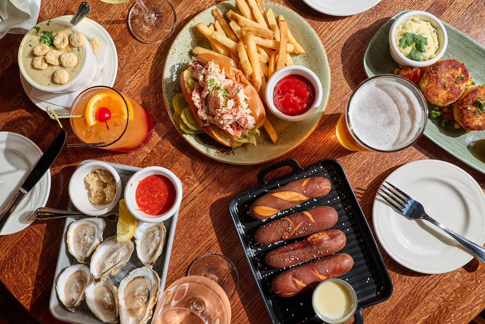 A variety of seafood, including raw oysters on the half shell and a bowl of creamy clam chowder, along with fries, a lobster roll with lobster salad, and various dipping sauces on a wooden table. There are also glasses of drinks, one with a lemon sli