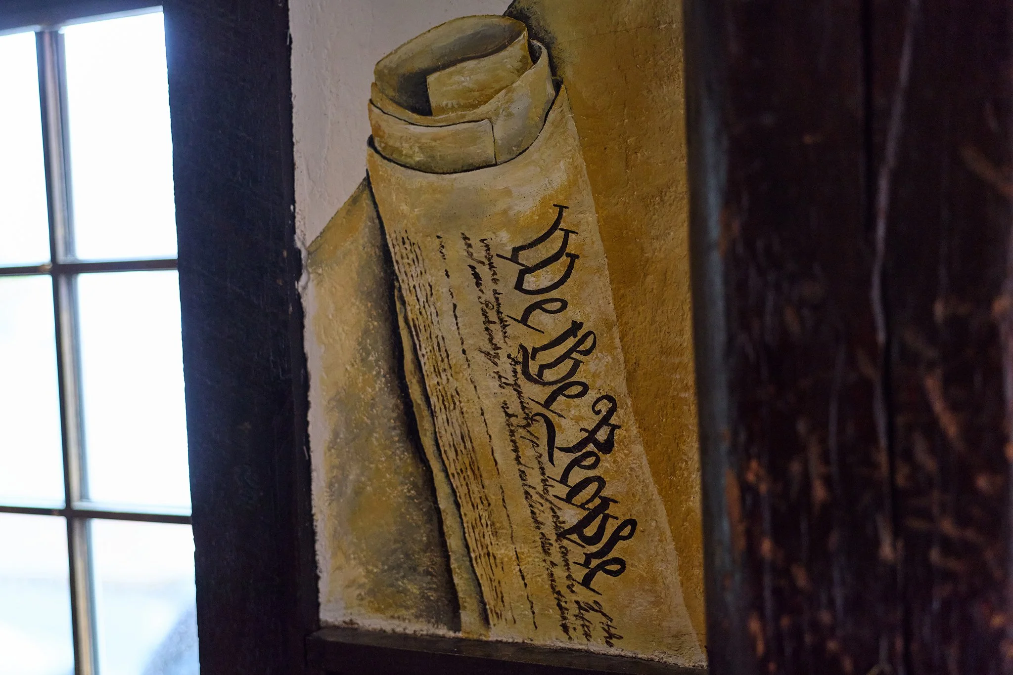 Close-up of an old, rolled parchment or manuscript with handwritten text, placed on a wooden surface. In the background, there is a window with a grid pattern letting in natural light.