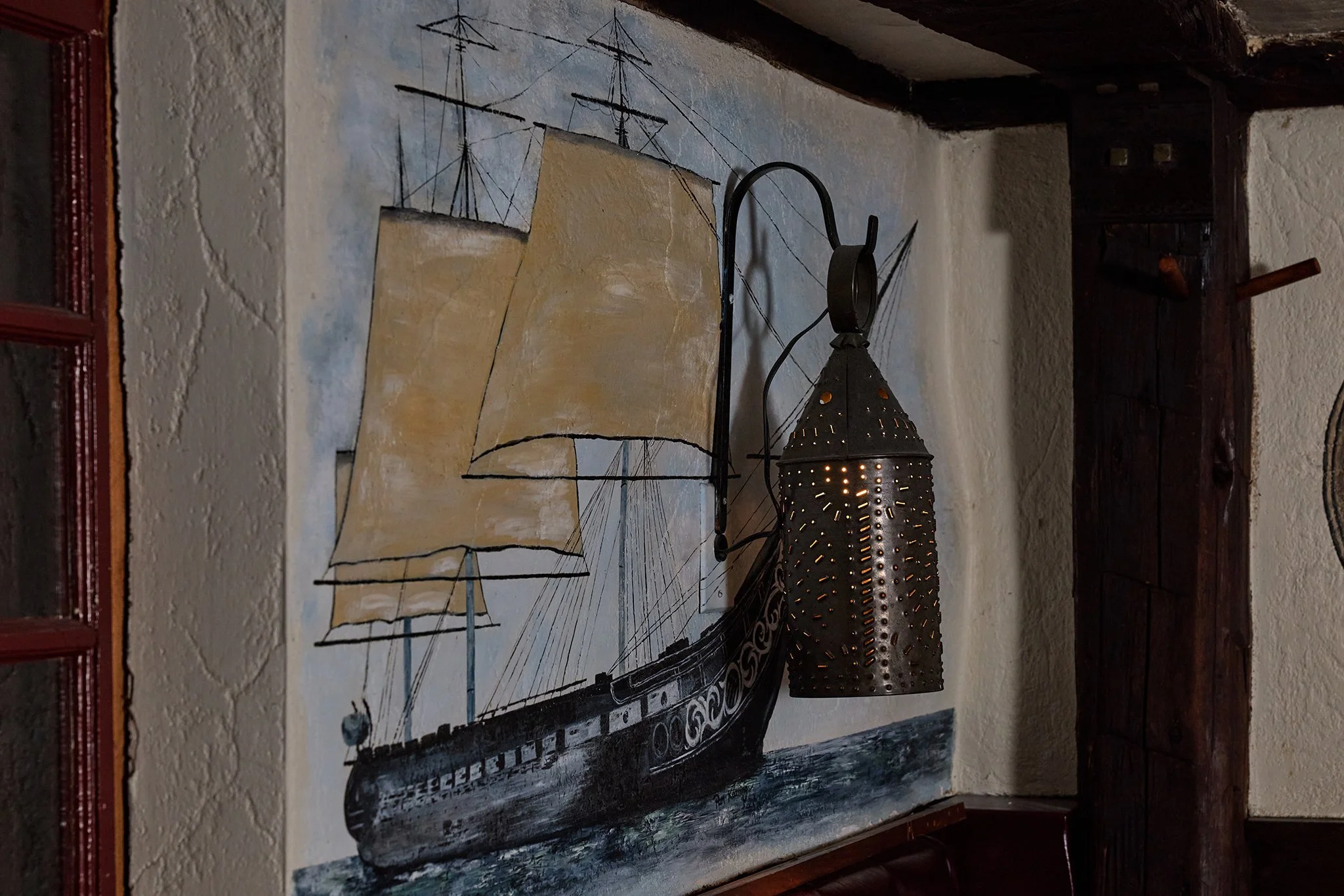 A painting of a large sailing ship with white sails, mounted on a wall behind a decorative metal lamp with perforations.