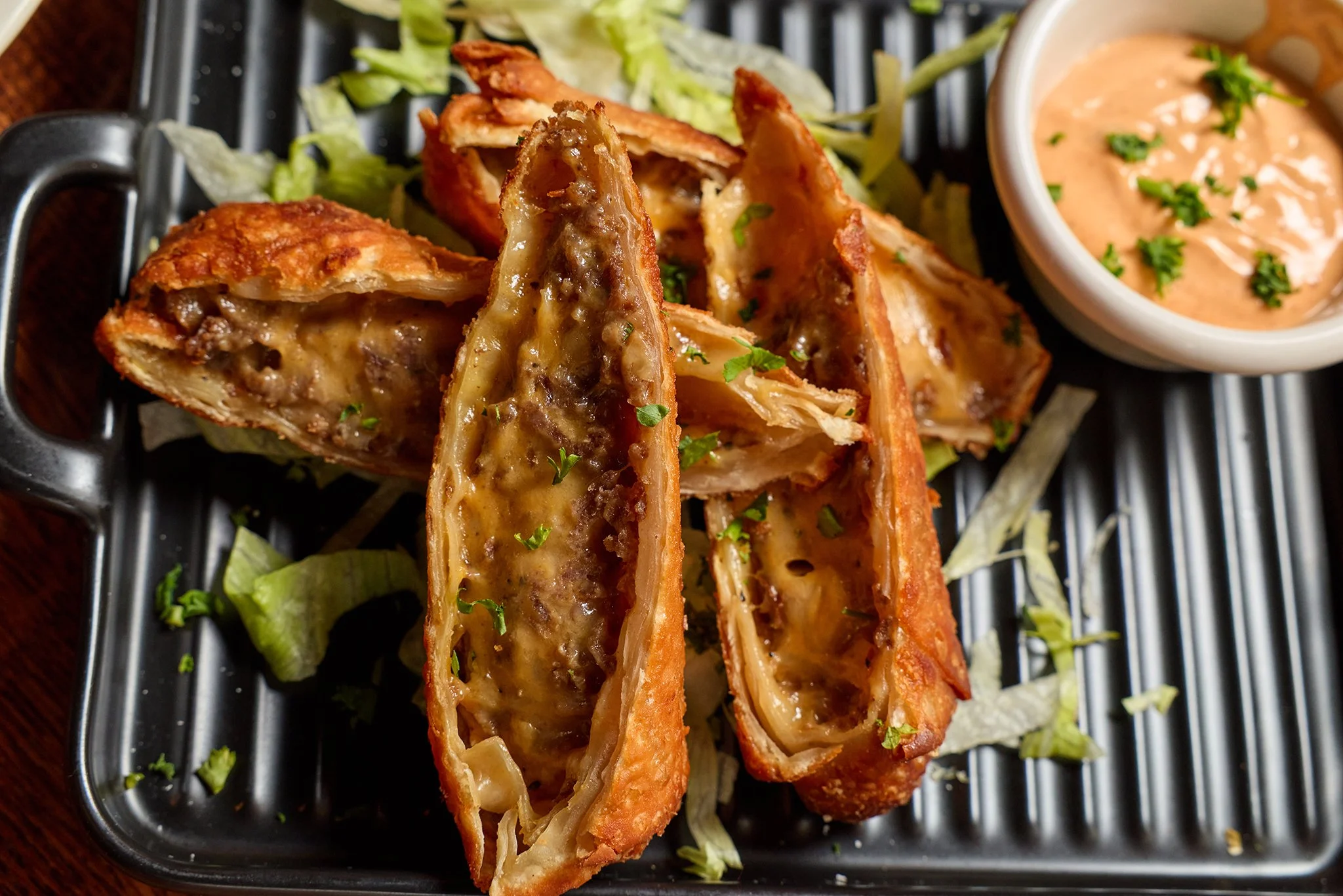 Fried spring rolls cut in half with savory filling, served on a bed of lettuce with a side of creamy dipping sauce garnished with chopped herbs.