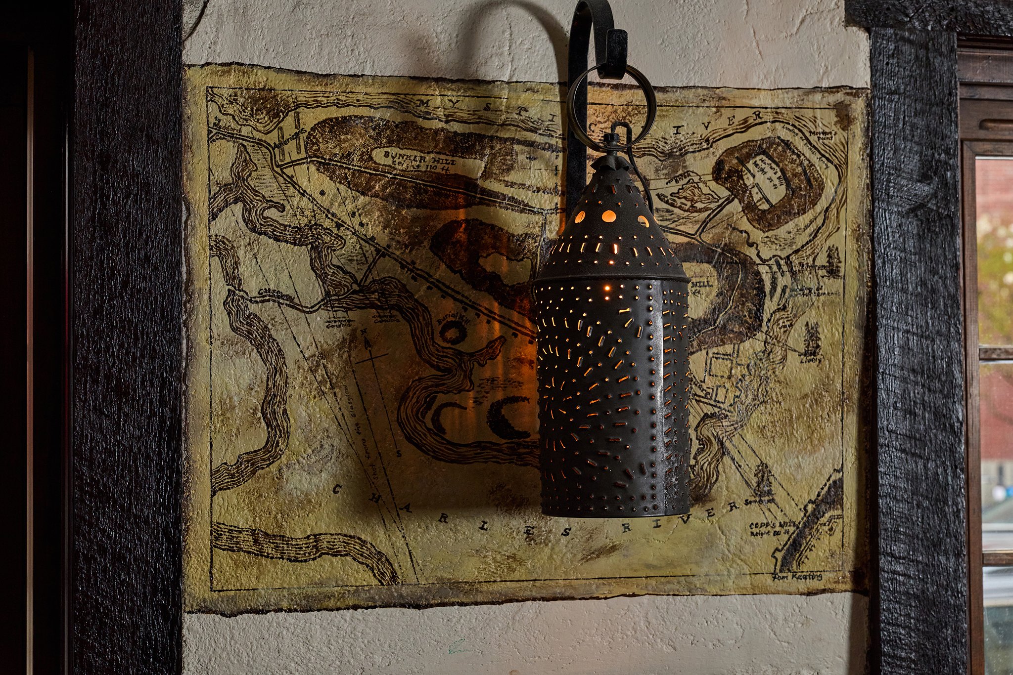 A vintage map of the river and surrounding area, mounted on a wall with black wooden beams framing it. A black perforated lantern hangs in front of the map, casting a warm glow.