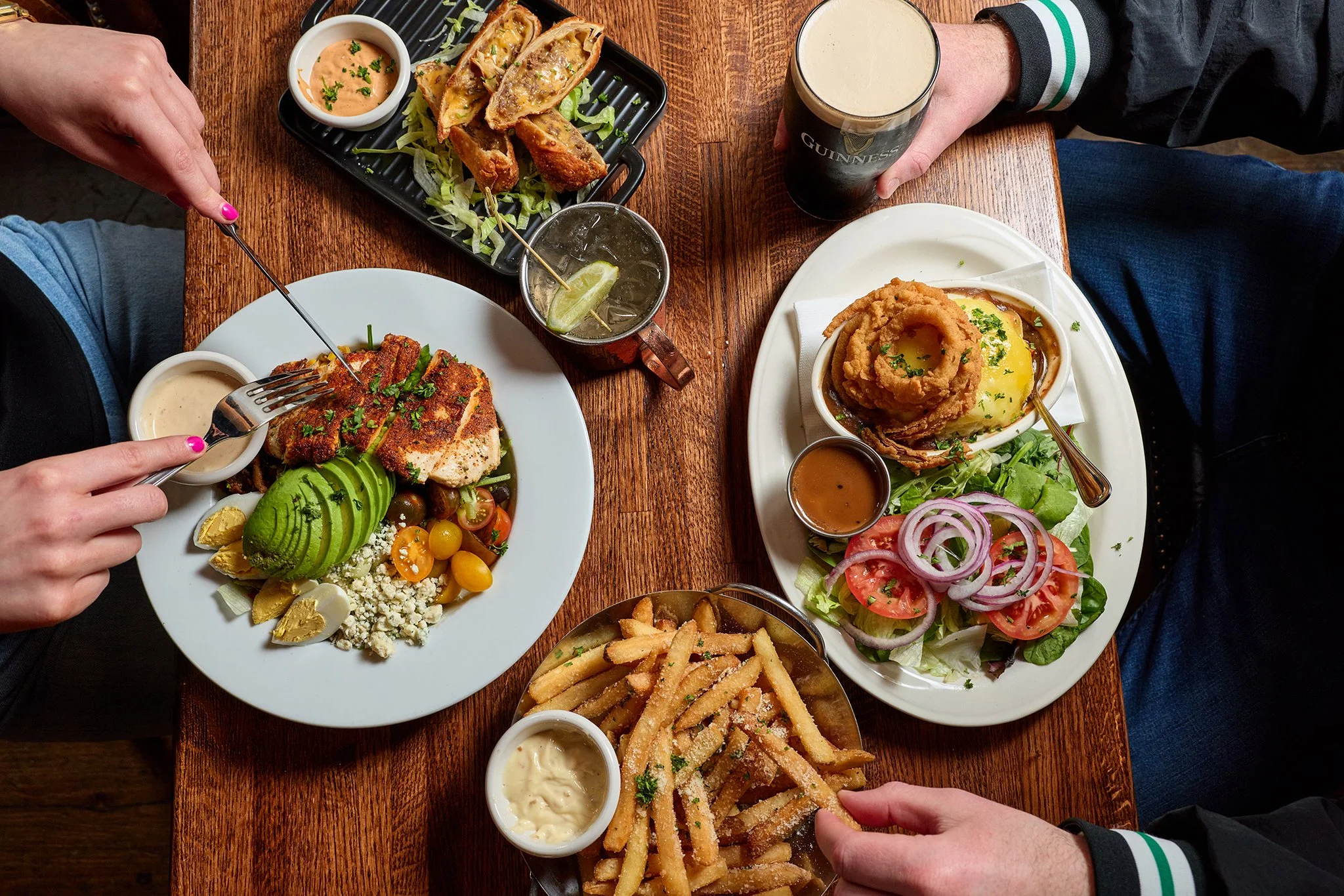A top-down view of a table with various dishes including a salad, fried foods with dipping sauces, and beverages, with two people reaching for the food.