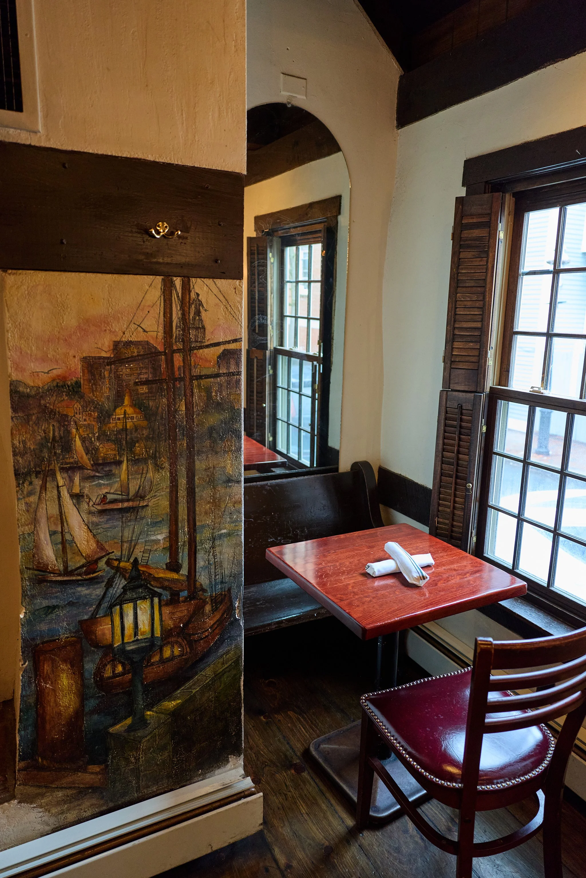 A cozy corner of a restaurant with a wooden table set with a cloth napkin and utensils, large windows with wooden shutters letting in natural light, and a wall painted with a maritime scene featuring a ship and buildings.
