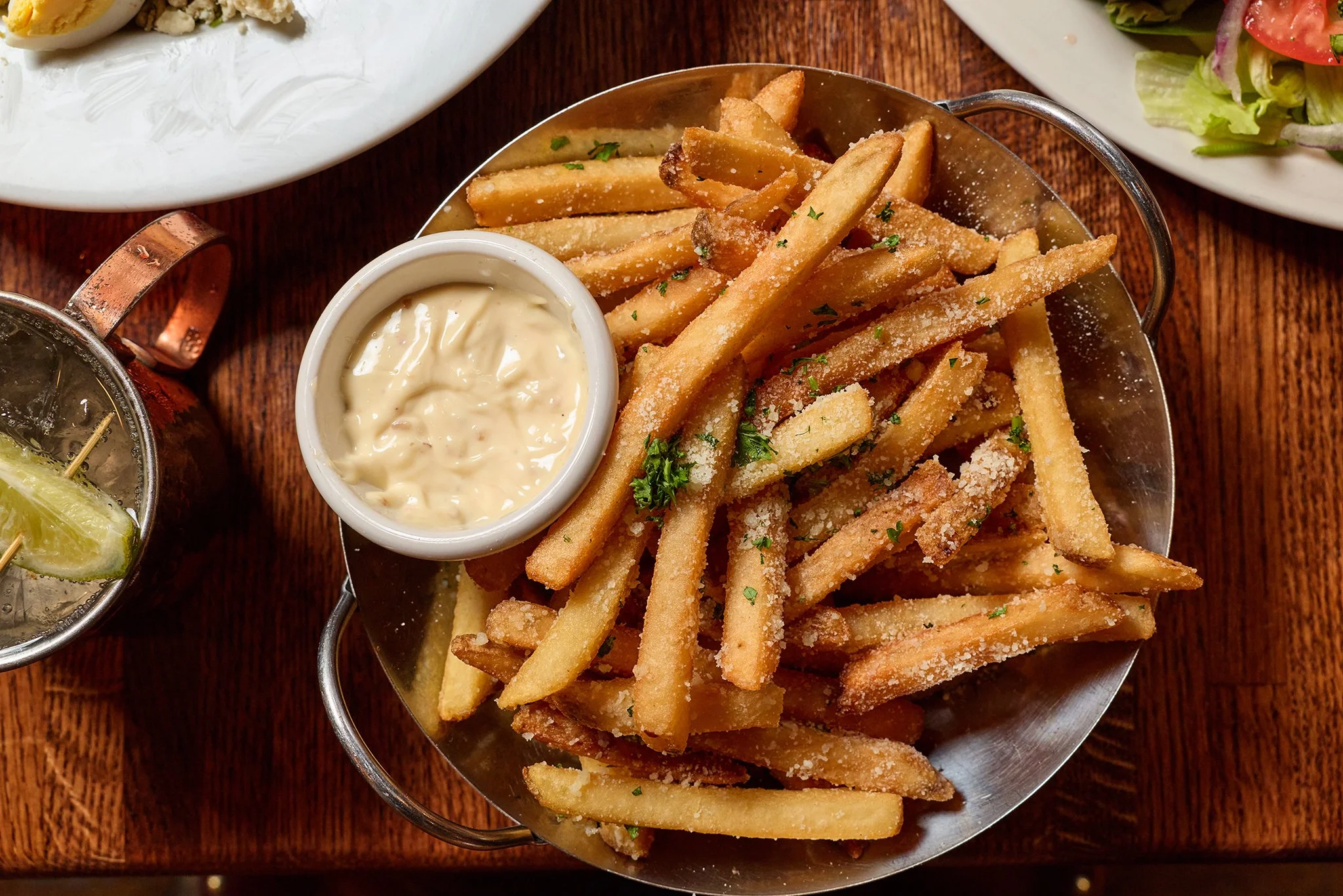 A basket of French fries garnished with parsley and sprinkled with salt. Served with a side of creamy dipping sauce. Part of a meal including salad and a drink on a wooden table.