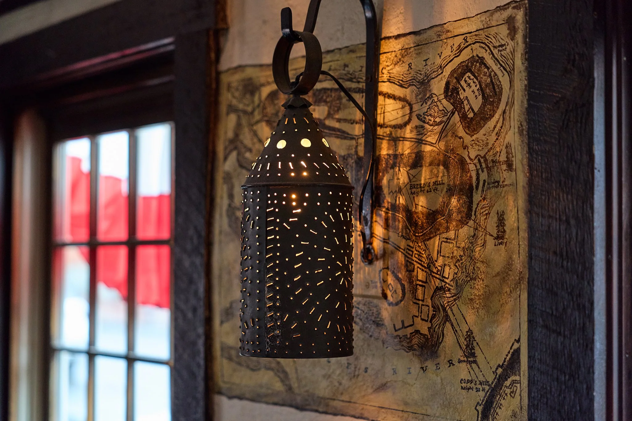 A decorative metal lantern with perforations hanging on a black iron bracket on a textured yellowish-brown wall. Behind the lantern, there is a map or painting with geographical markings. On the left side, a window with red curtains is partially visi