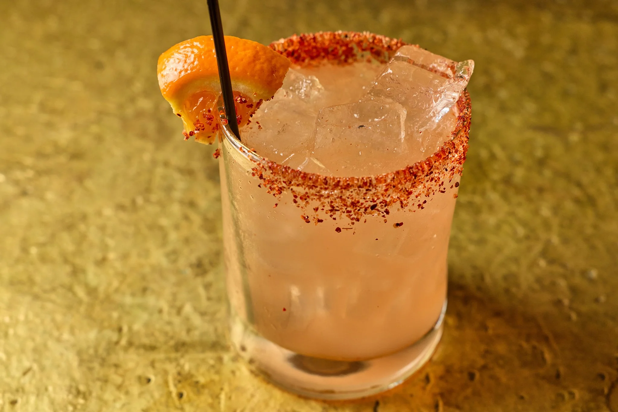 A pink cocktail drink in a tall glass with salted rim, garnished with a slice of orange and a black straw, on a yellowish countertop.