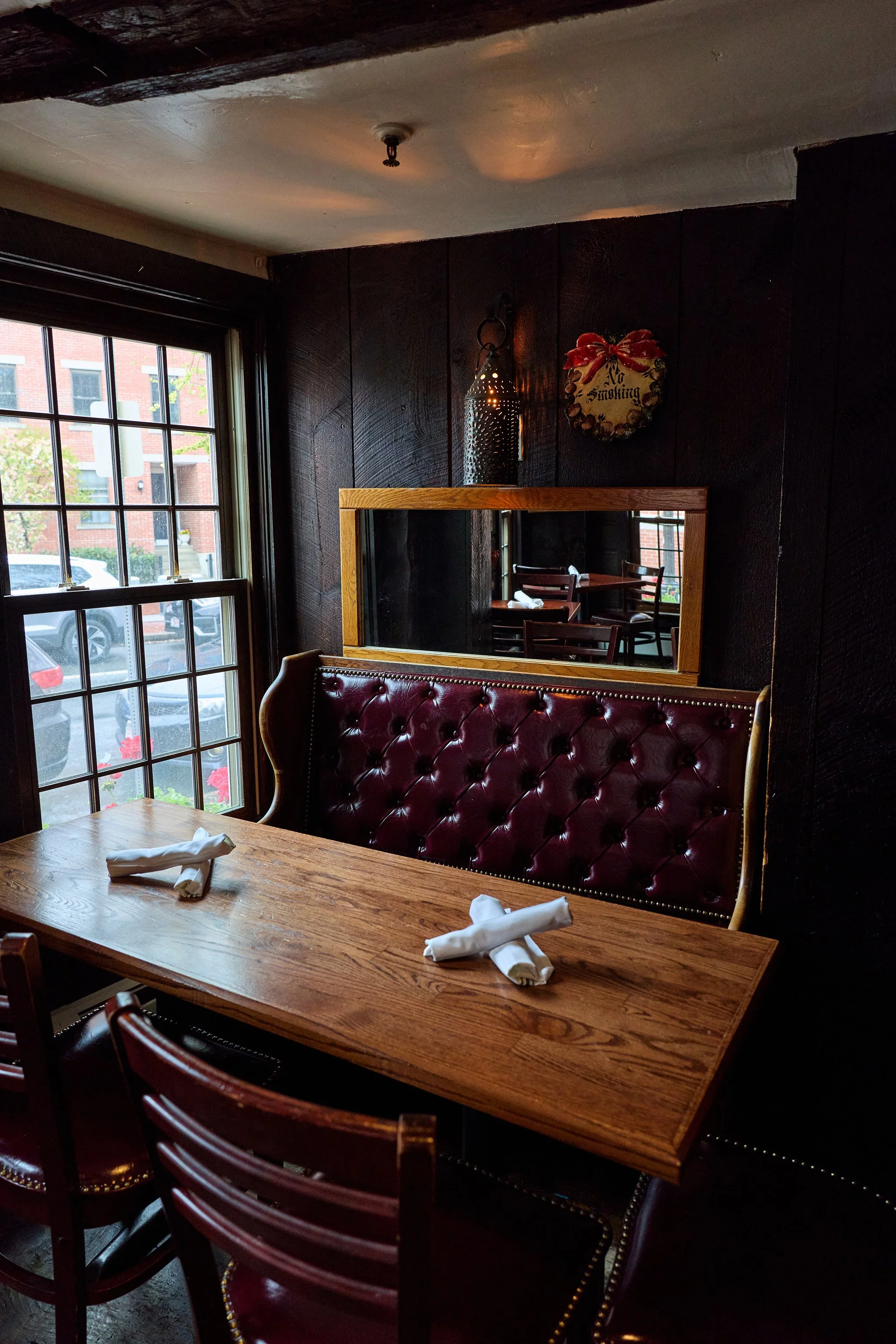 Interior of a cozy restaurant with a wooden table set with white napkins. A red leather upholstered booth with nailhead trim is against a dark wood paneled wall, which features a large mirror and a hanging decorative lantern. A window with grid panes shows parked cars and a brick building outside.