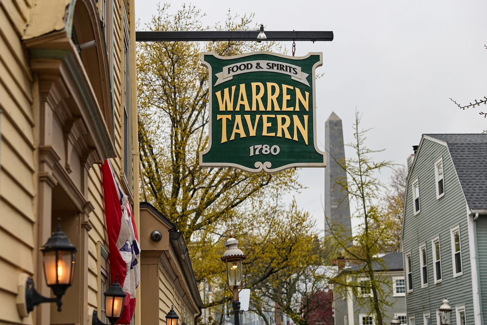 A green sign hanging from a building reads "Food & Spirits Warren Tavern 1780." In the background, trees with yellow leaves, neighboring houses, and a tall chimney are visible, with some black lantern-style lights mounted on the building's exterior.