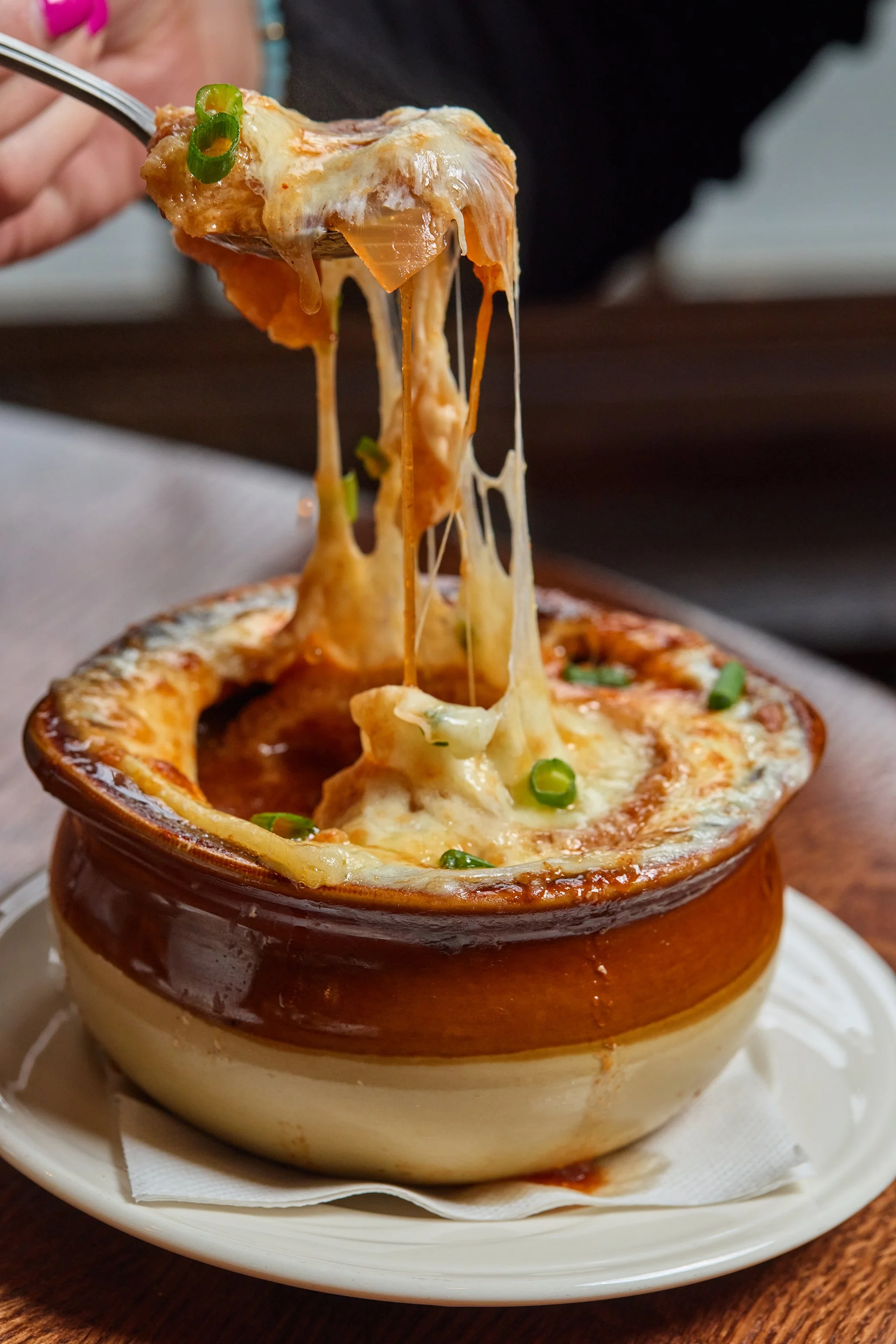 A baked dish of cheesy casserole with melted cheese and chopped green onions in a brown ceramic bowl on a white plate with a paper napkin underneath.