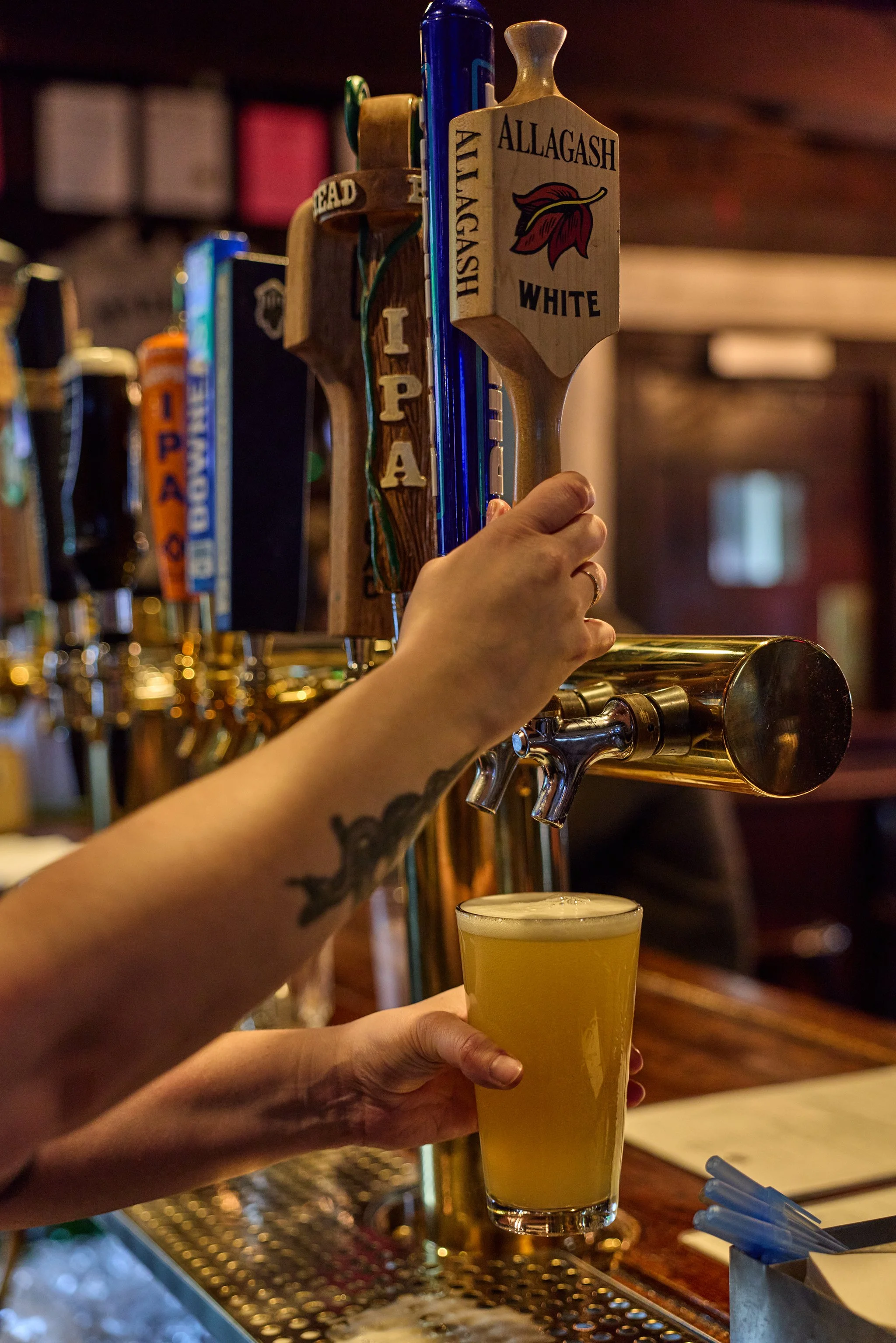 A person's hand with a tattoo is holding a pint of hazy yellow beer at a bar. In the background, another person's hand is pouring draft beer from a tap labeled 'Allagash White' with a logo of a red leaf. The bar has a polished wooden counter and a me