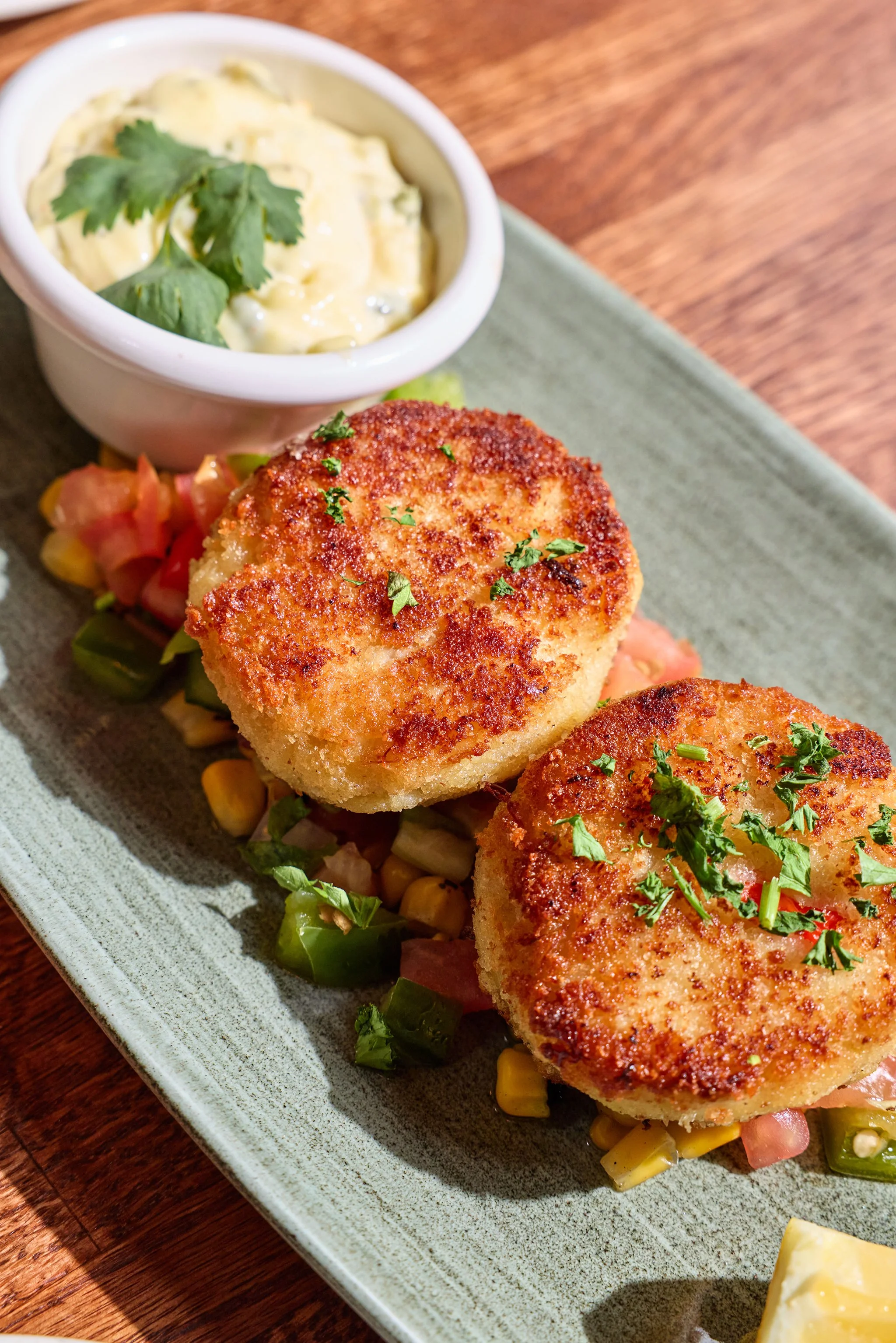 Close-up of breaded fish fillets garnished with chopped herbs, served on a plate with corn, diced tomatoes, green vegetables, and a side of creamy dip garnished with cilantro.