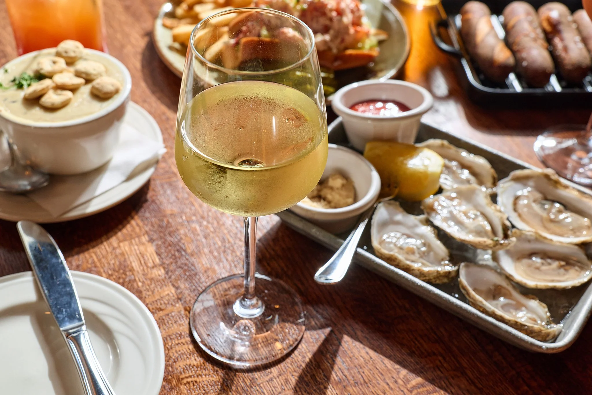 A glass of white wine on a wooden table surrounded by various dishes including oysters on a tray, a bowl of chowder, a plate of ribs, and small containers of sauces and lemon slices.