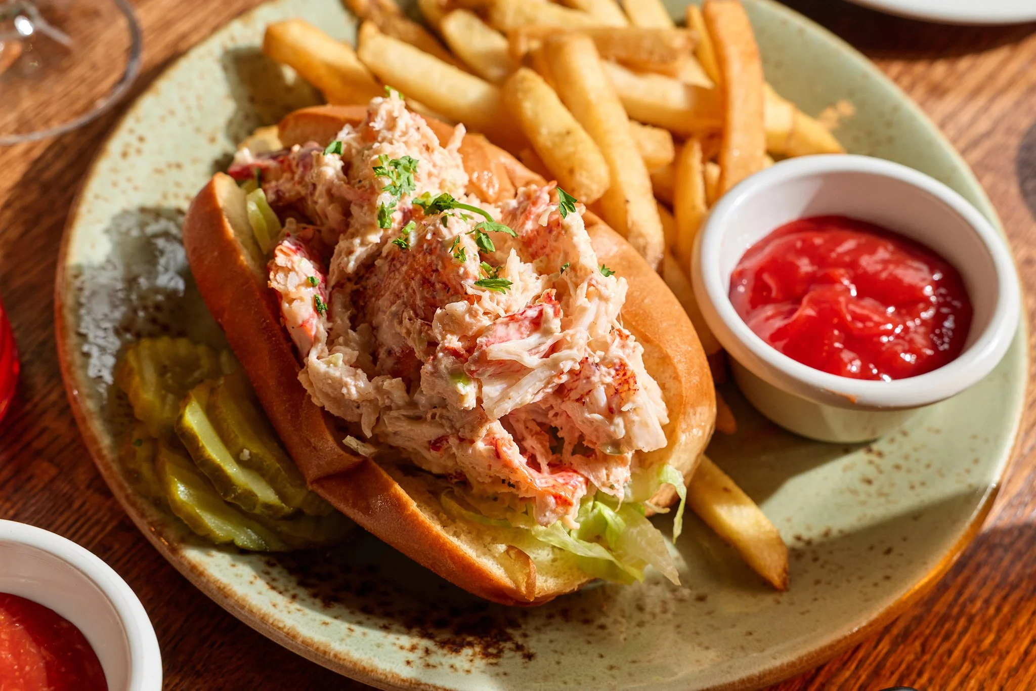 Lobster roll sandwich with lettuce in a bun, served with French fries and ketchup in a small white dish.