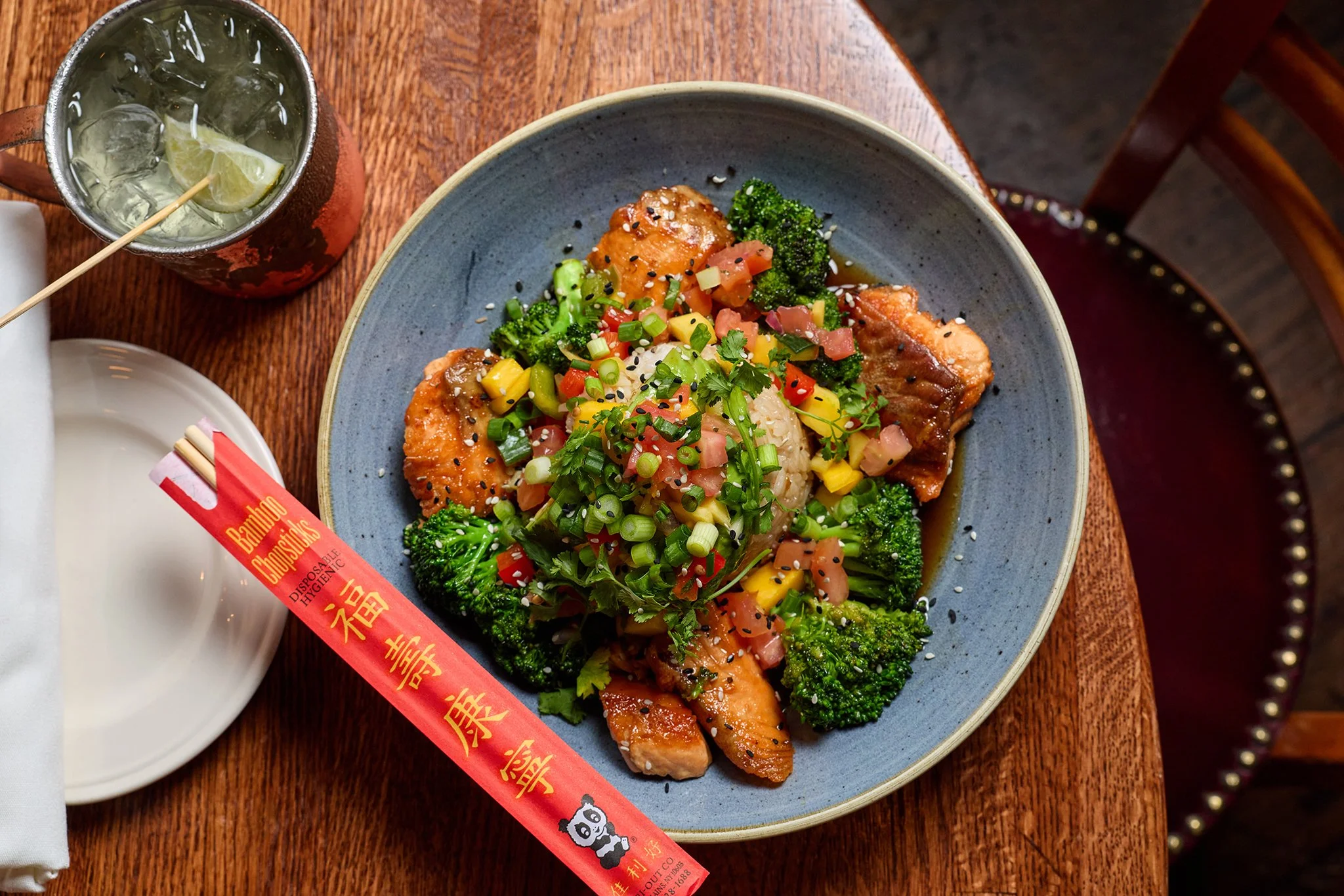 A bowl of Asian-style stir-fry with broccoli, diced vegetables, and sesame seeds, with a drink containing lemon and ice, and a pair of red chopsticks with gold Chinese characters on a napkin, all on a wooden table.