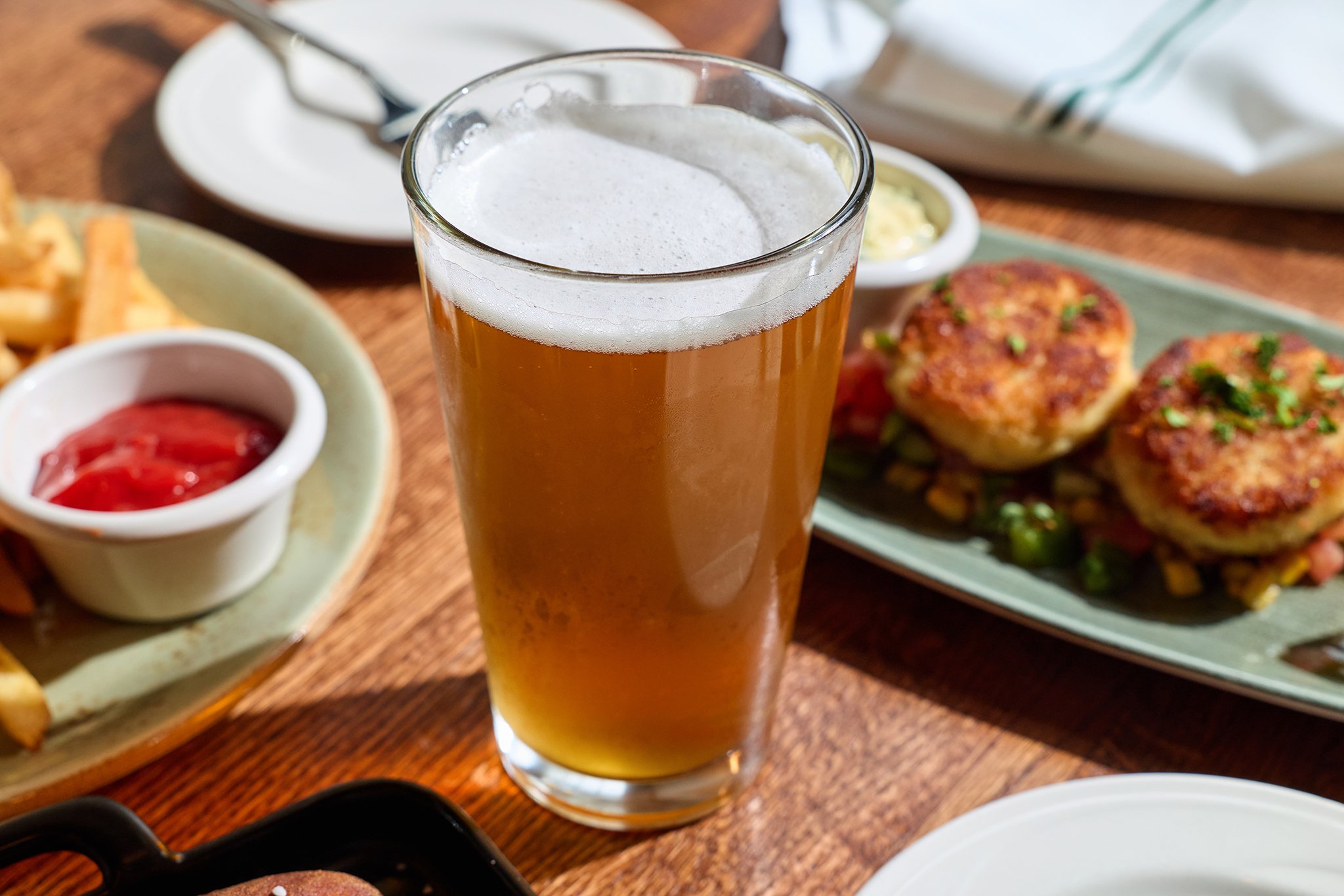 A glass of beer on a wooden table with various plates of food, including what appears to be meatballs, fries, and dipping sauces in the background.