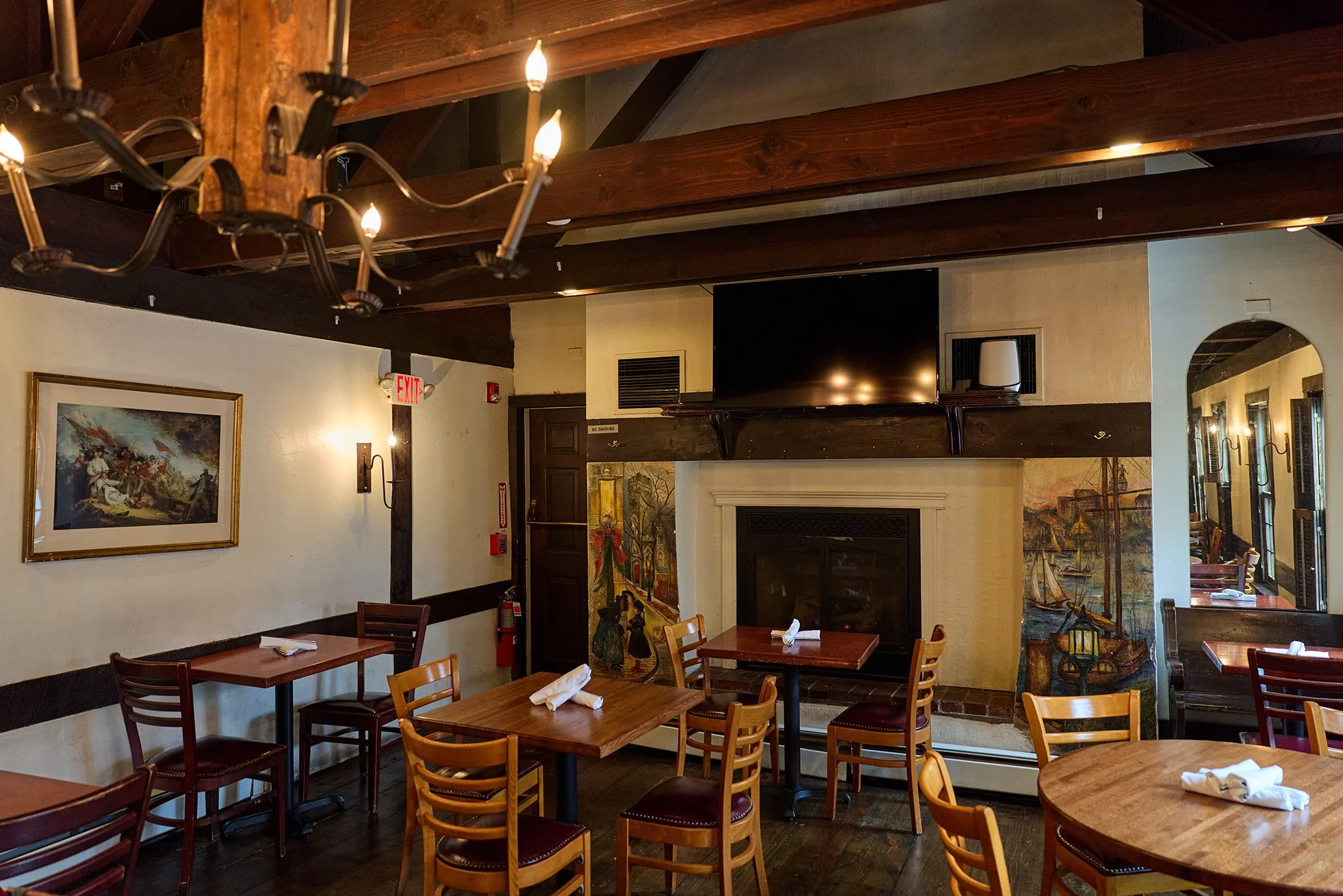 Interior of a cozy restaurant or café with wooden beams, framed paintings on the wall, tables set with napkins, and warm lighting.