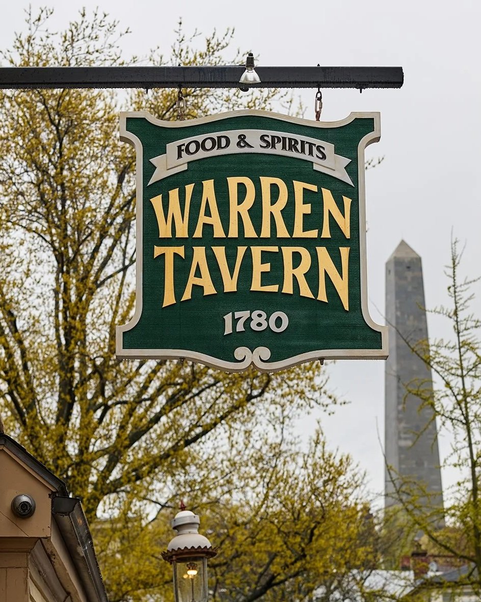 A sign for Warren Tavern, established in 1780, advertising food and spirits, hanging outside on a black pole against a background of trees with yellow leaves and a tall obelisk in the distance.