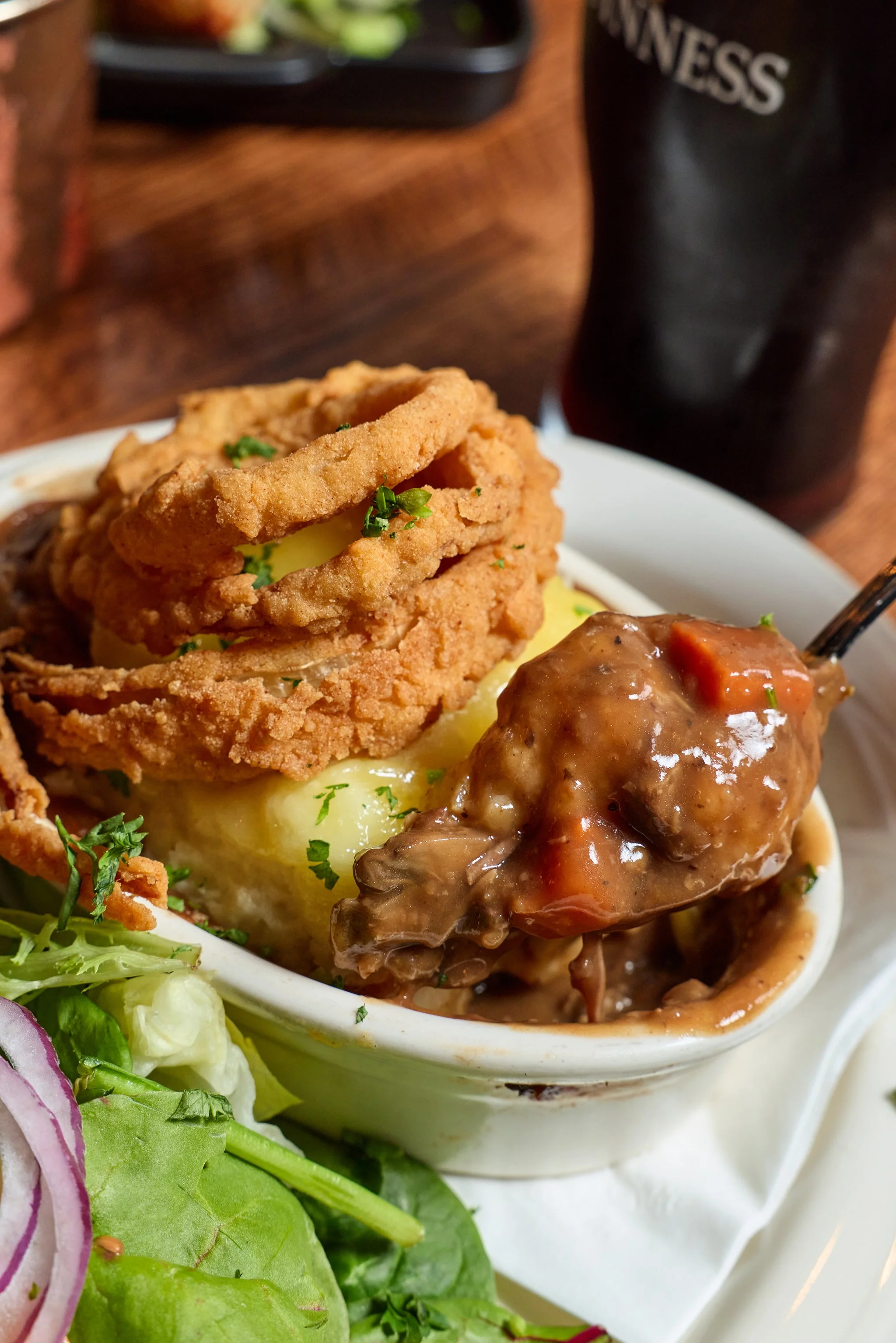 Close-up of a dish featuring mashed potatoes topped with fried onions and gravy, served with a side salad including lettuce, radish slices, and greens, in a white dish, with a dark drink container in the background.